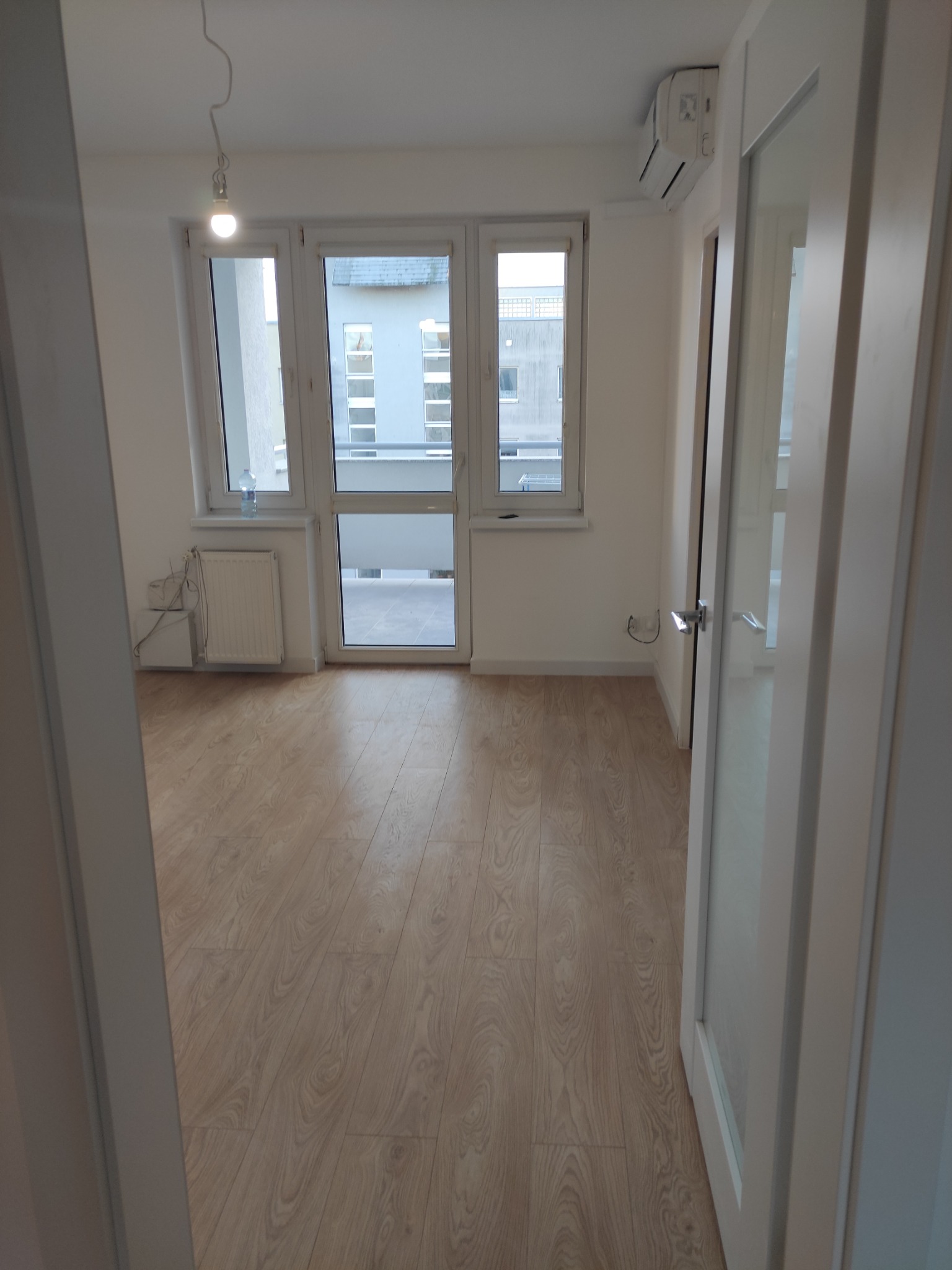 Jasny pokój po remoncie z panelami podłogowymi w odcieniach drewna, widok przez otwarte białe drzwi, grzejnik pod oknem i klimatyzator na ścianie.