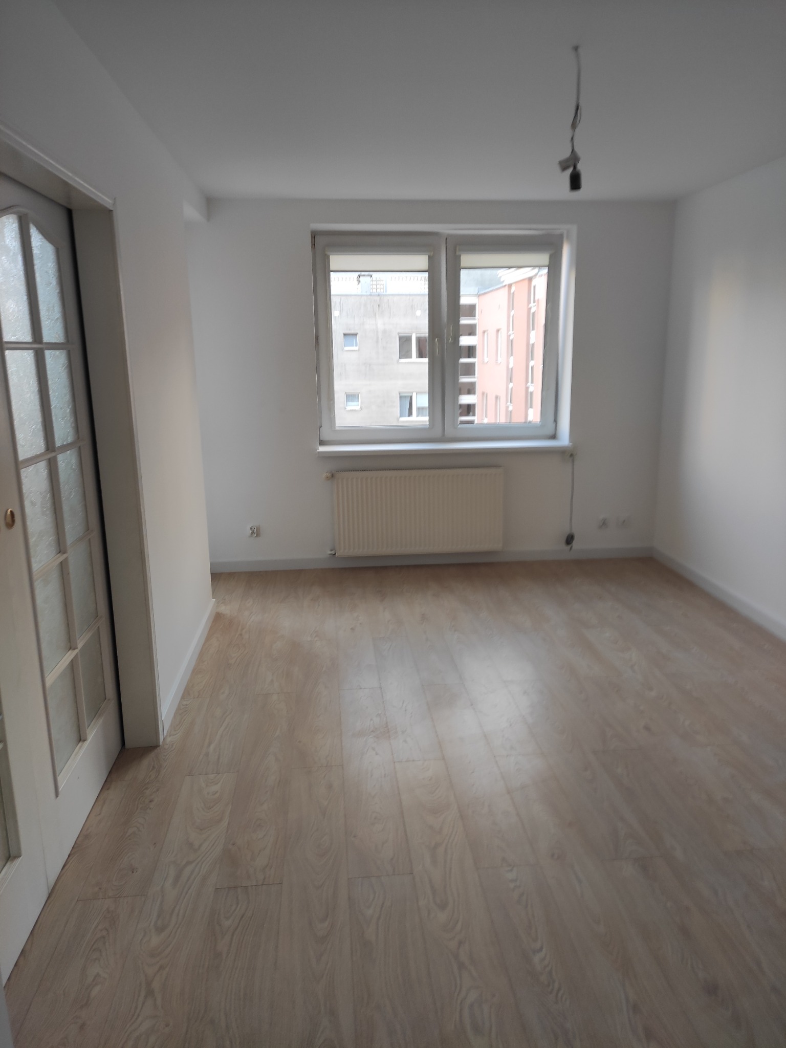 Pusty pokój z nową podłogą z paneli laminowanych, białe ściany, okno z widokiem na budynki, białe drzwi z przeszkleniami po lewej stronie.