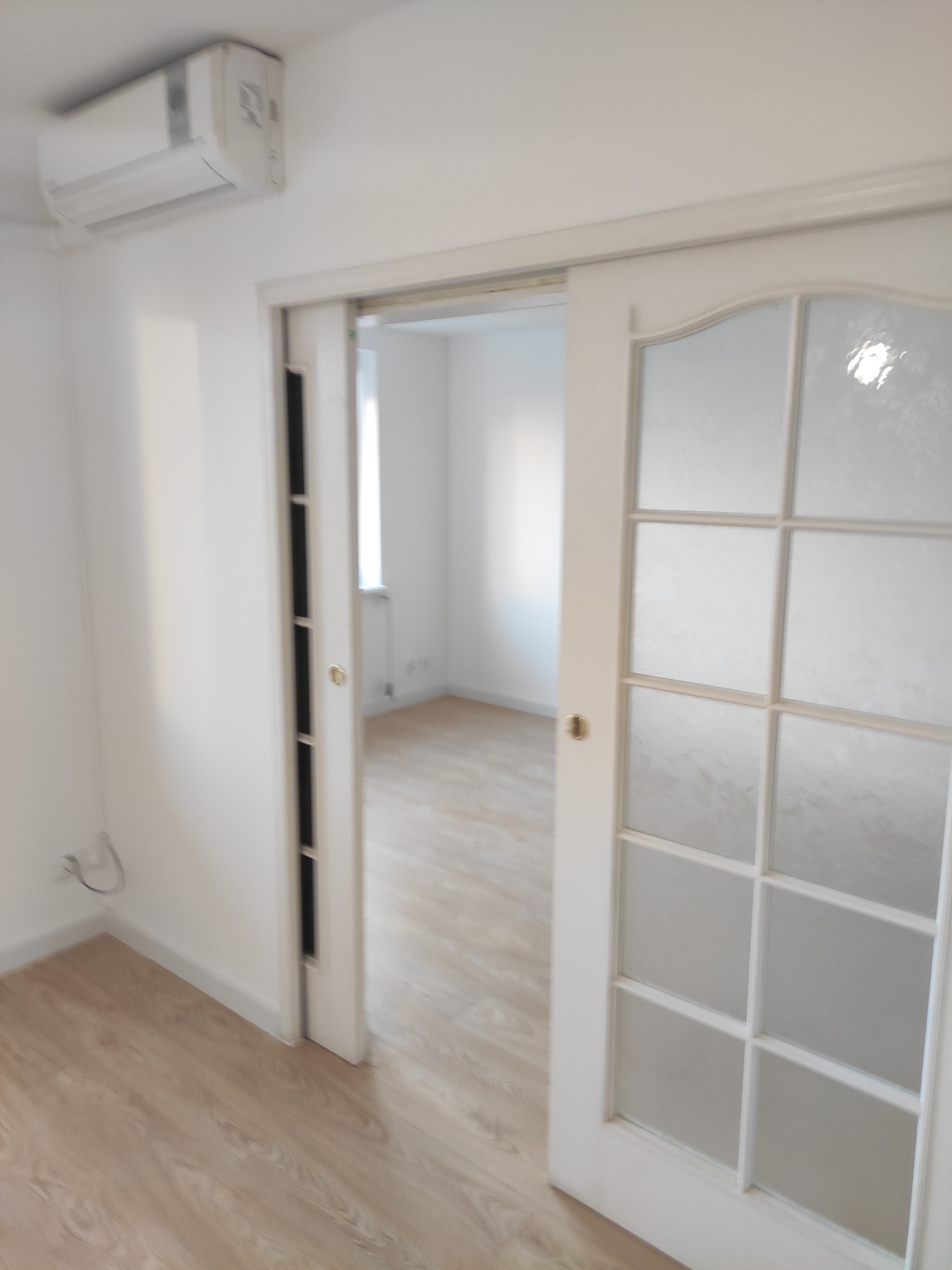 Białe przesuwne drzwi z matowego szkła, otwarte w jasnym pokoju z panelami podłogowymi i klimatyzatorem na ścianie.