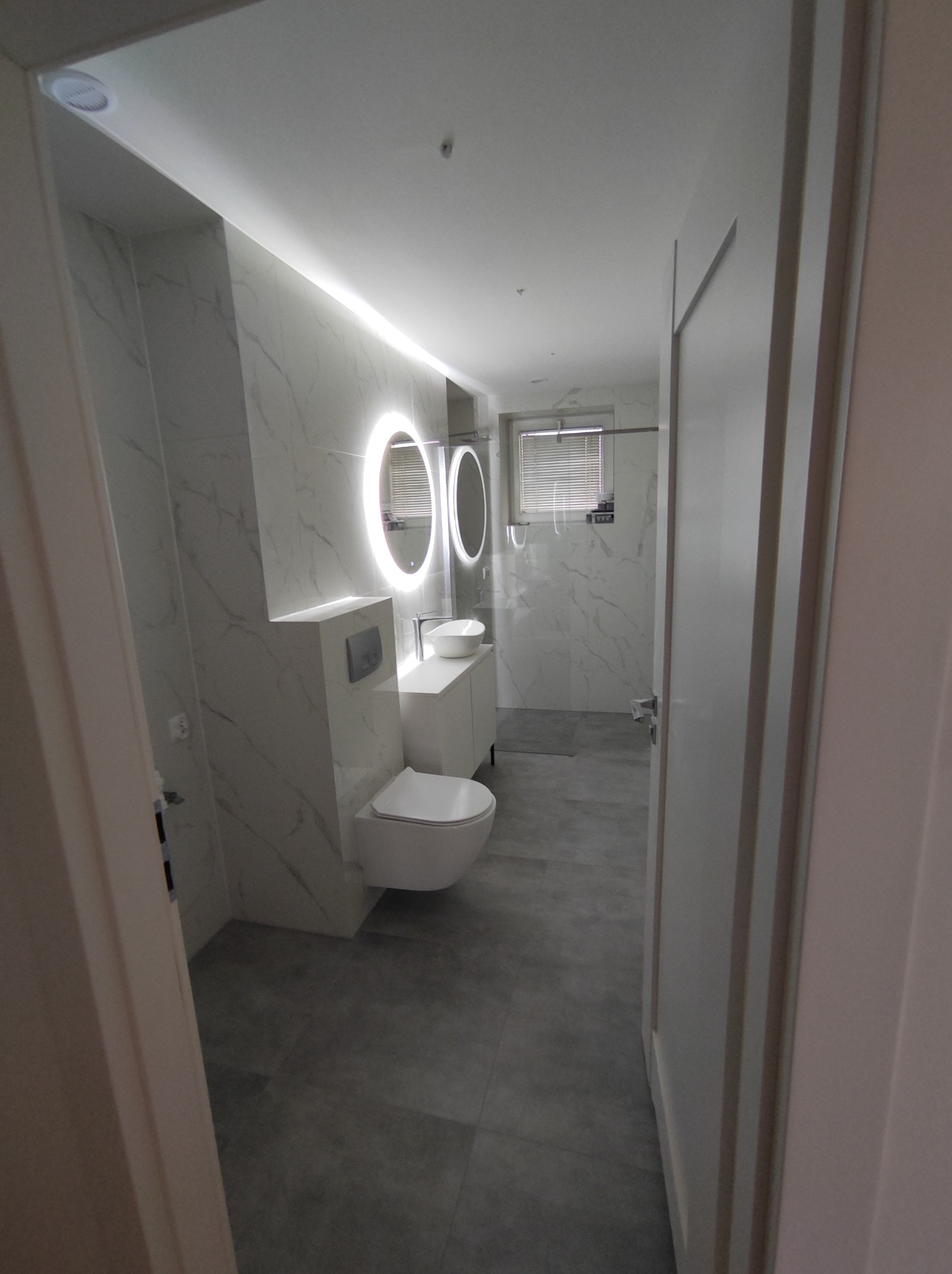 Nowoczesna łazienka z podświetlanymi okrągłymi lustrami, wiszącą toaletą i szarymi płytkami imitującymi marmur, widziana zza uchylonych drzwi.