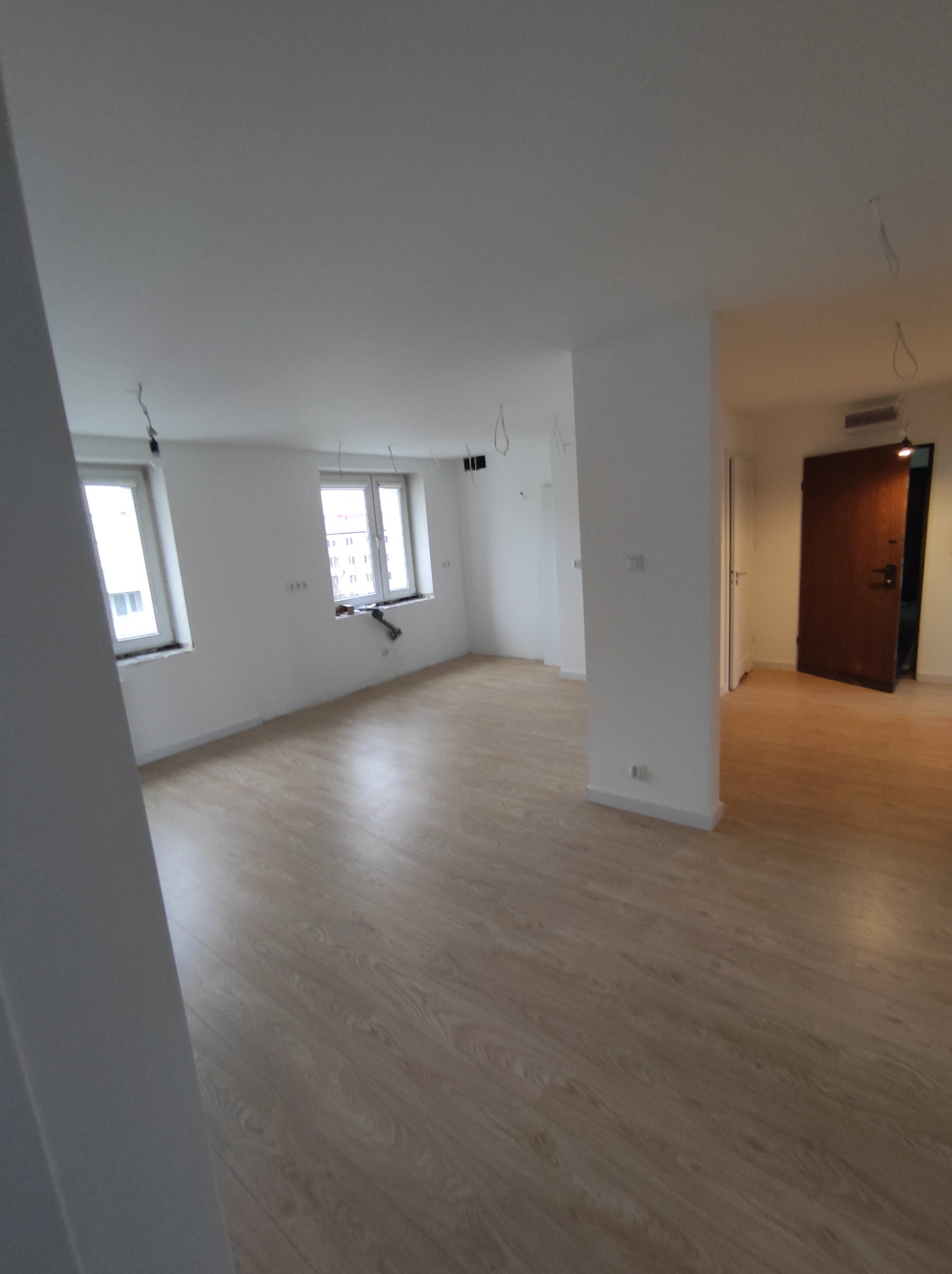 Wnętrze mieszkania w trakcie prac wykończeniowych: świeżo położone panele podłogowe o wzorze drewna, białe ściany z widocznymi przewodami elektrycznymi, okna z widokiem na budynki oraz ciemne drzwi...