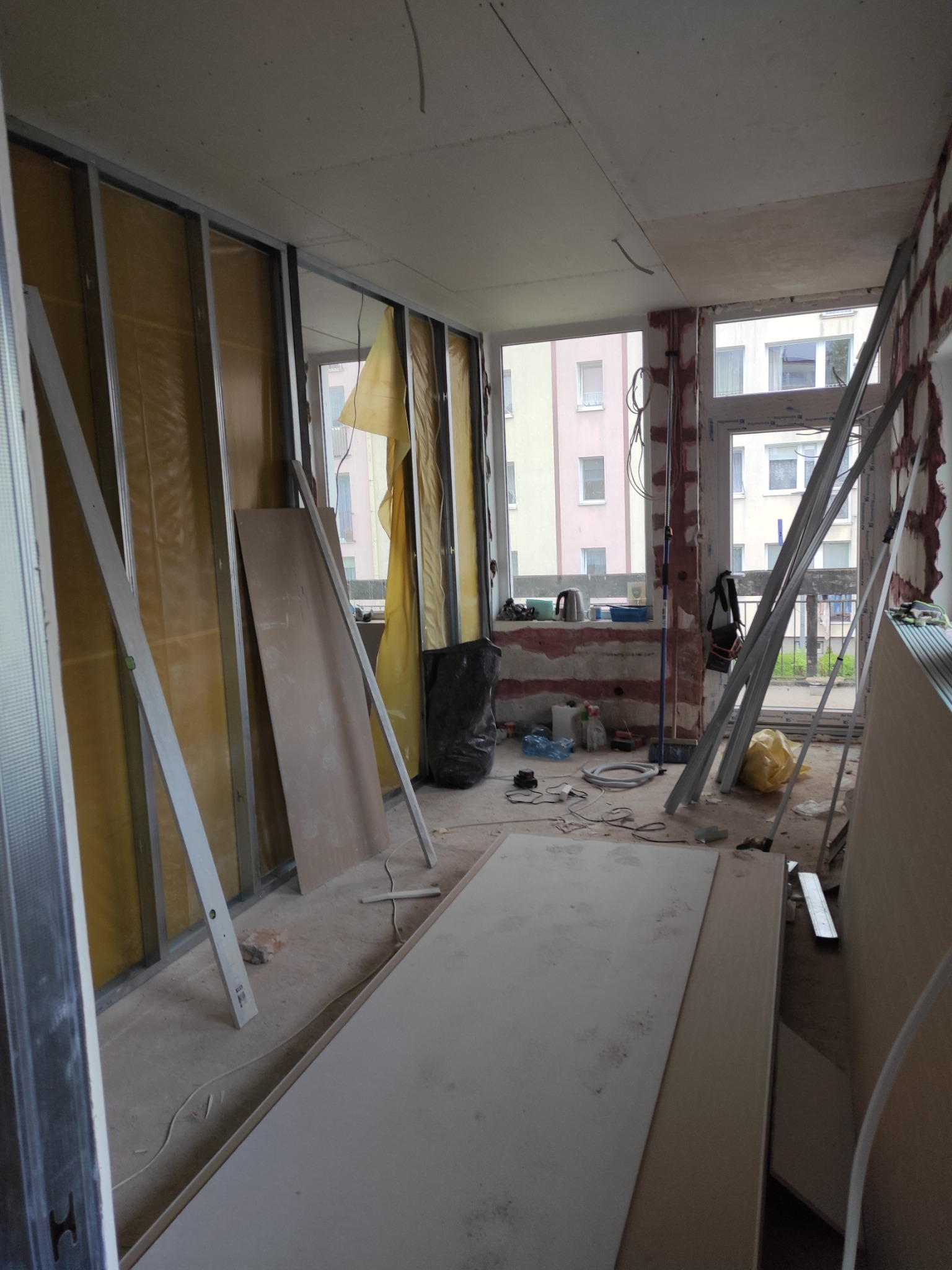 Wnętrze w trakcie prac: metalowa konstrukcja ścian działowych z wypełnieniem z wełny mineralnej, płyty gipsowo-kartonowe oparte o ścianę, widoczne przewody elektryczne i narzędzia na podłodze, okno...
