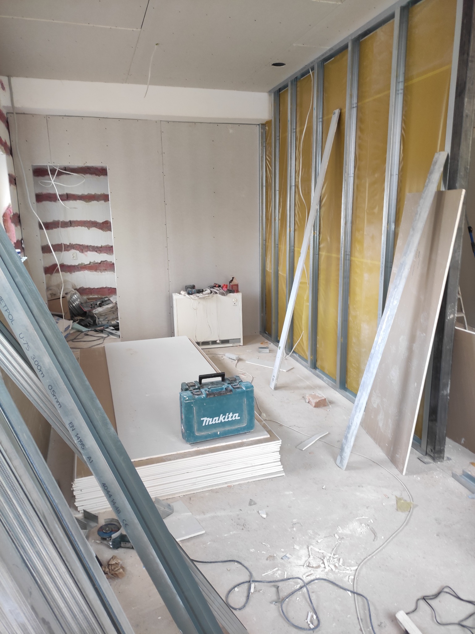 Montaż ścian działowych z płyt karton gips z widoczną izolacją, profile stalowe, narzędzia Makita i stos płyt gipsowych w surowym wnętrzu.