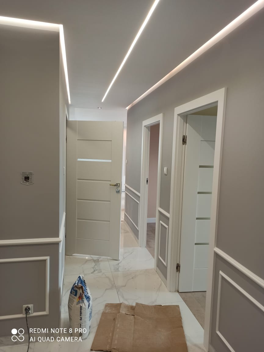 Szare wnętrze korytarza z białymi panelami dekoracyjnymi, drzwiami wewnętrznymi i ledowym oświetleniem sufitowym; na marmurowej podłodze leży worek zaprawy i karton zabezpieczający.