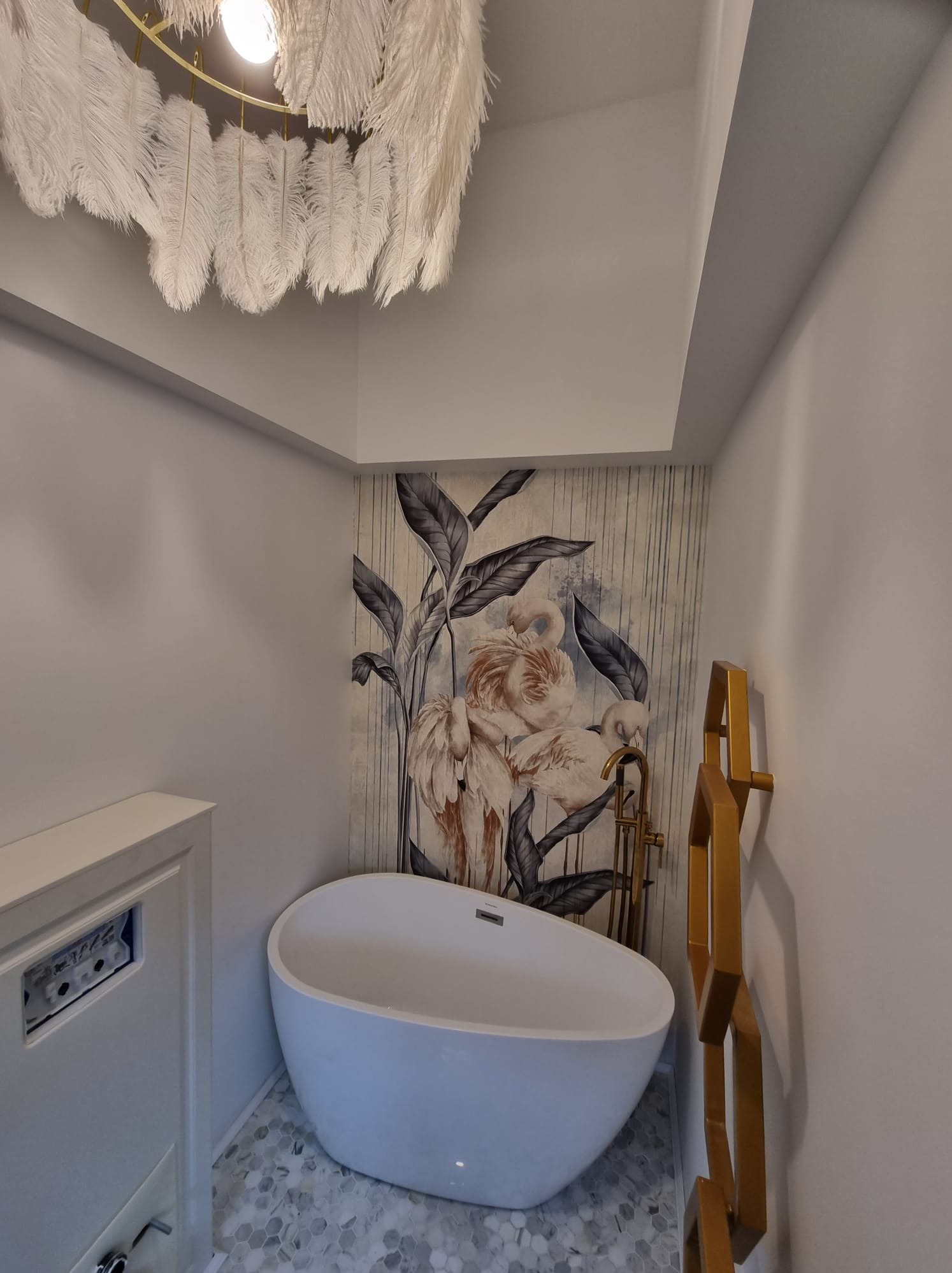 Nowoczesna łazienka z wolnostojącą białą wanną, złotą armaturą i grzejnikiem dekoracyjnym w kształcie heksagonów. Na ścianie tapeta z motywem flamingów i tropikalnych liści. Sufit zdobi designerski...