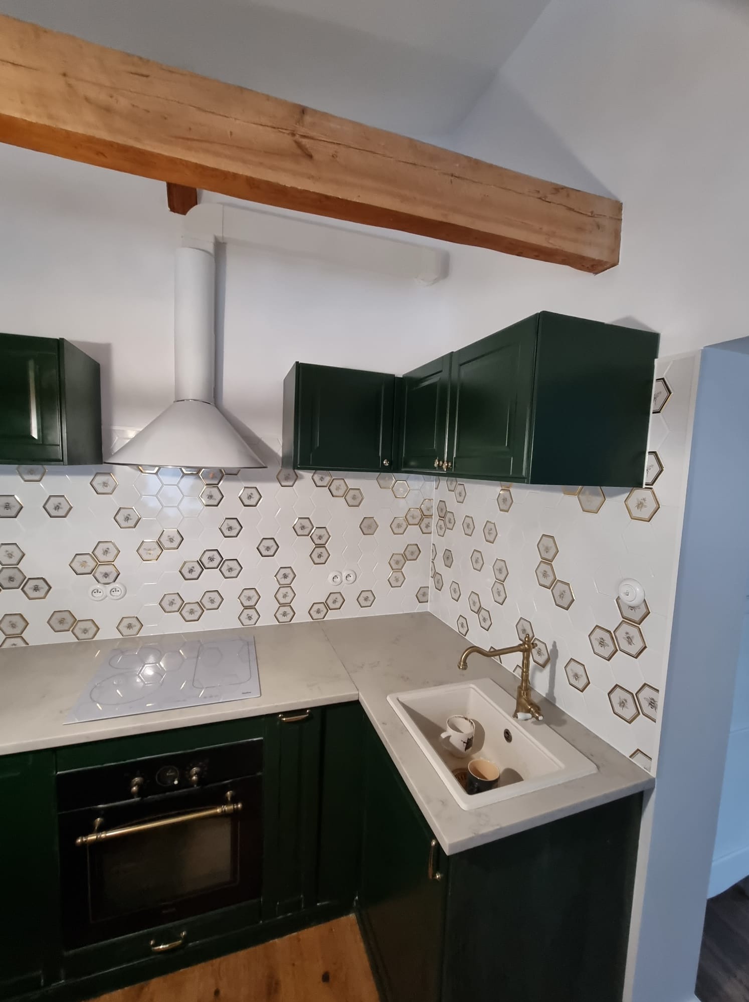 Wykończona kuchnia z ciemnozielonymi szafkami, białym blatem, wzorzystymi heksagonalnymi płytkami z motywem pszczół i złotą armaturą, widoczna drewniana belka stropowa.