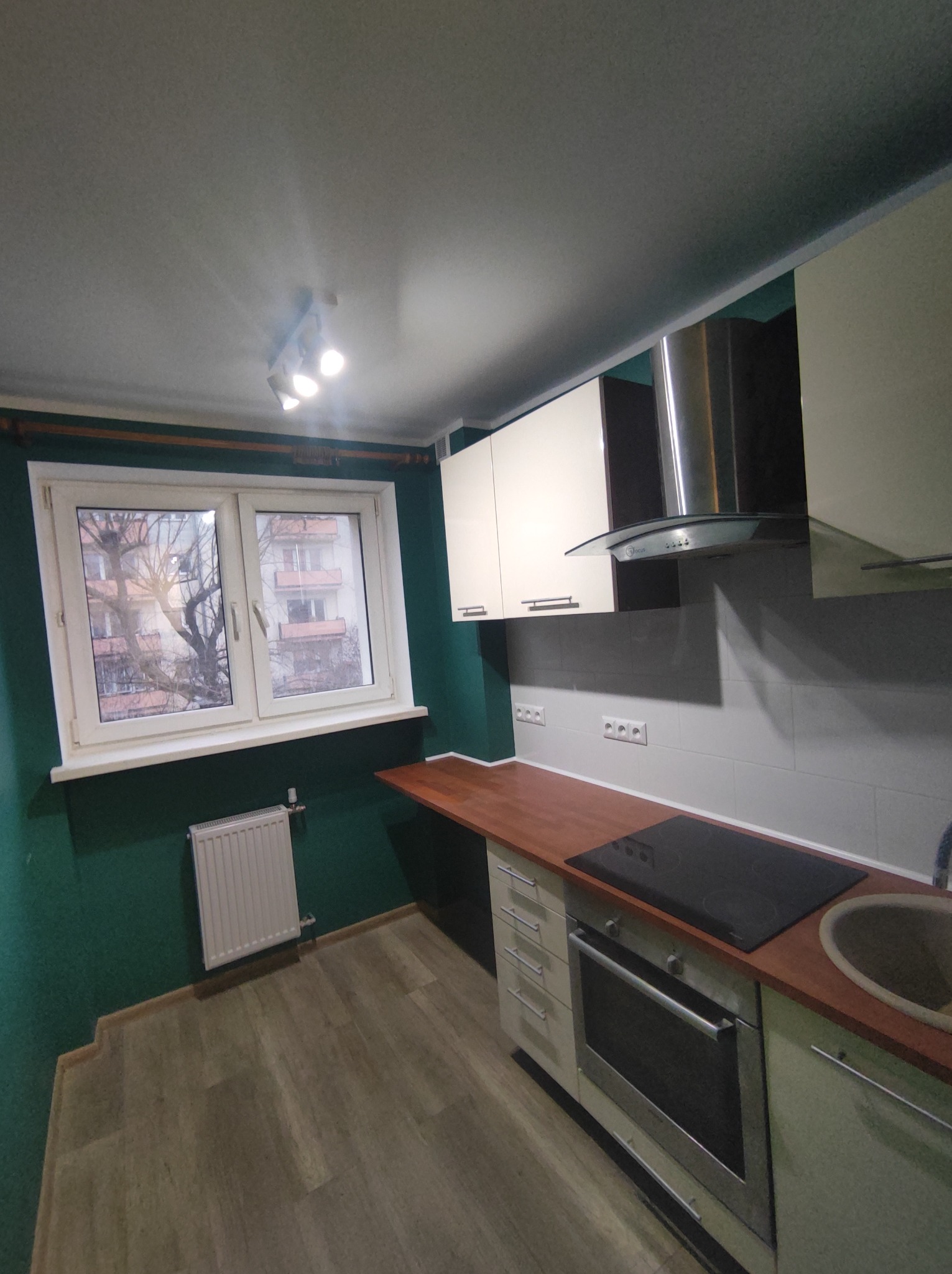 Wnętrze kuchni po remoncie: zielone ściany, jasne szafki, drewniany blat, okno z widokiem na budynek mieszkalny, grzejnik pod oknem.