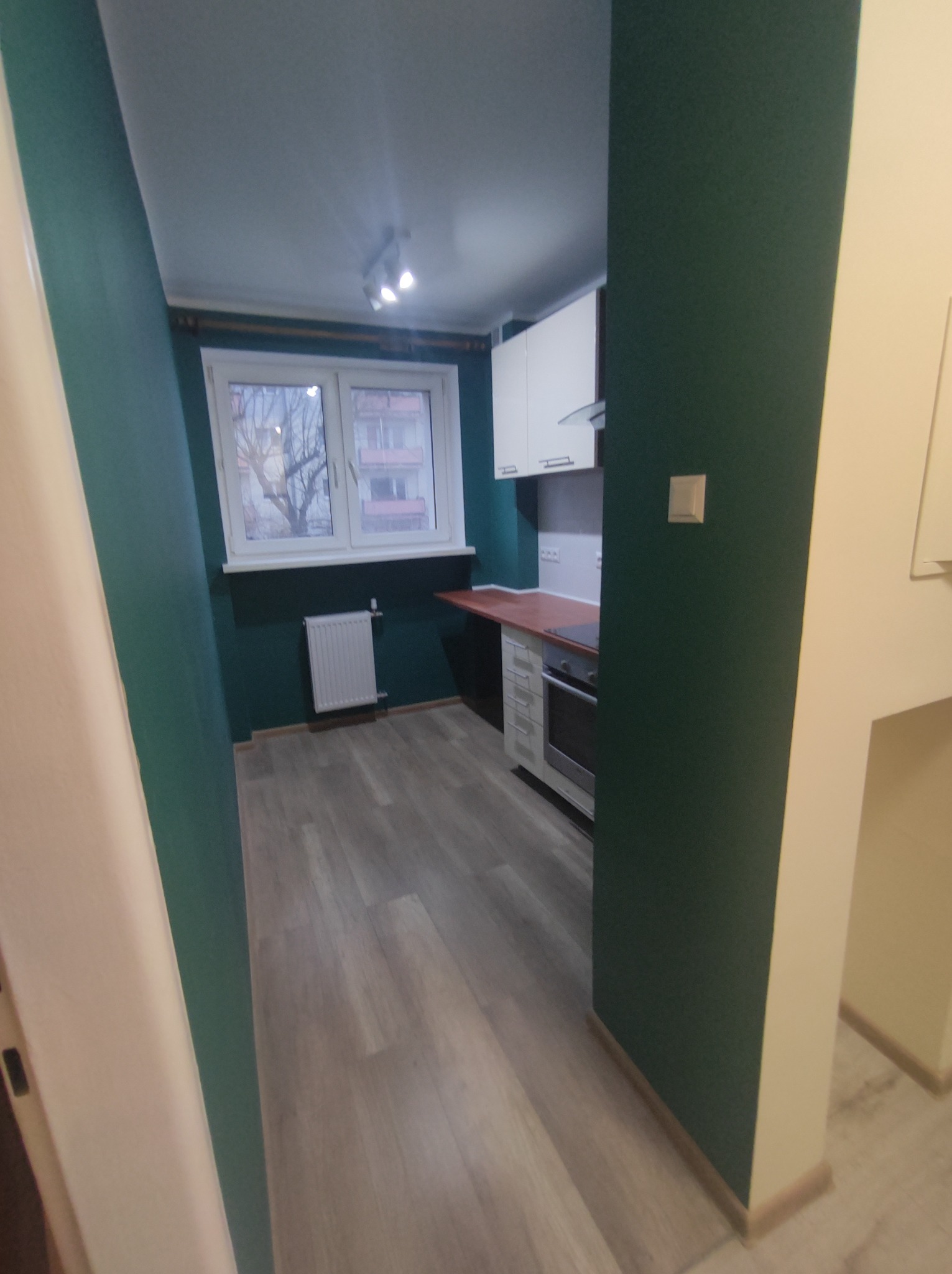 Wykończona kuchnia w wąskim pomieszczeniu z zielonymi ścianami, jasnymi panelami podłogowymi i białymi szafkami, widok na okno z grzejnikiem.