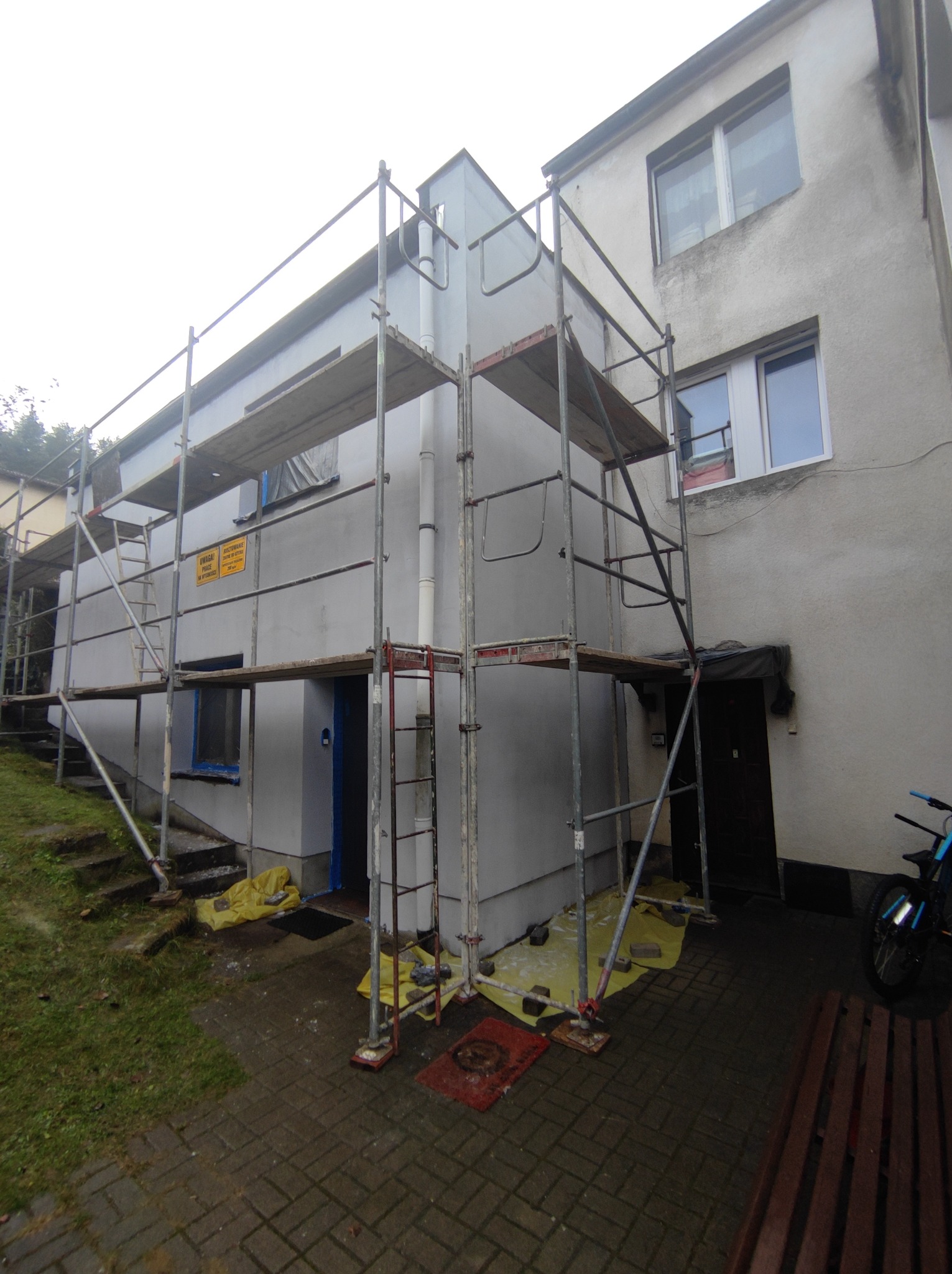 Budynek mieszkalny w trakcie odświeżania elewacji, z rozstawionymi rusztowaniami, zabezpieczonymi oknami i żółtą folią ochronną na ziemi.