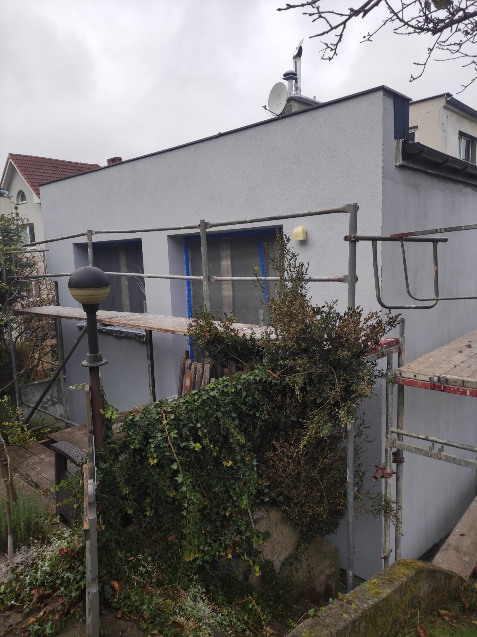 Świeżo pomalowany, szary budynek z rusztowaniem i zabezpieczonymi oknami, widok z zarośniętego ogrodu w pochmurny dzień.