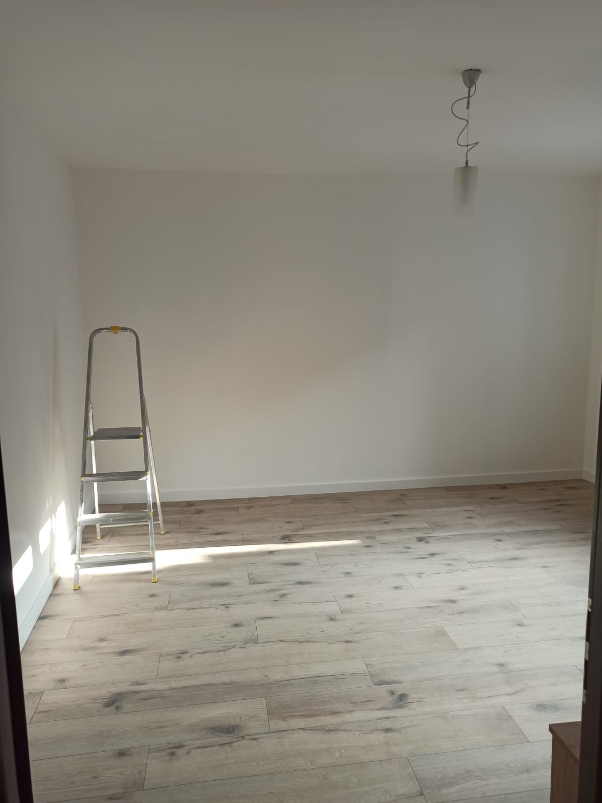 Wnętrze pokoju po remoncie: świeżo pomalowane białe ściany, ułożone jasne panele podłogowe imitujące drewno, aluminiowa drabina oparta o ścianę, widoczny fragment sufitu z lampą.