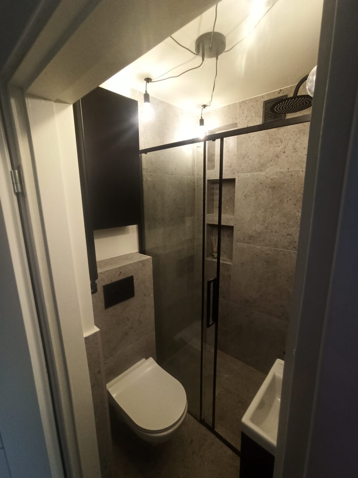 Nowoczesna łazienka z kabiną prysznicową typu walk-in, wiszącą toaletą i umywalką nablatową, wykończona płytkami imitującymi kamień, oświetlona lampami wiszącymi z żarówkami Edison.