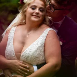 Uśmiechnięta panna młoda w koronkowej sukni z kwiatami we włosach, obejmowana przez pana młodego w okularach i bordowej muszce, który ją całuje.