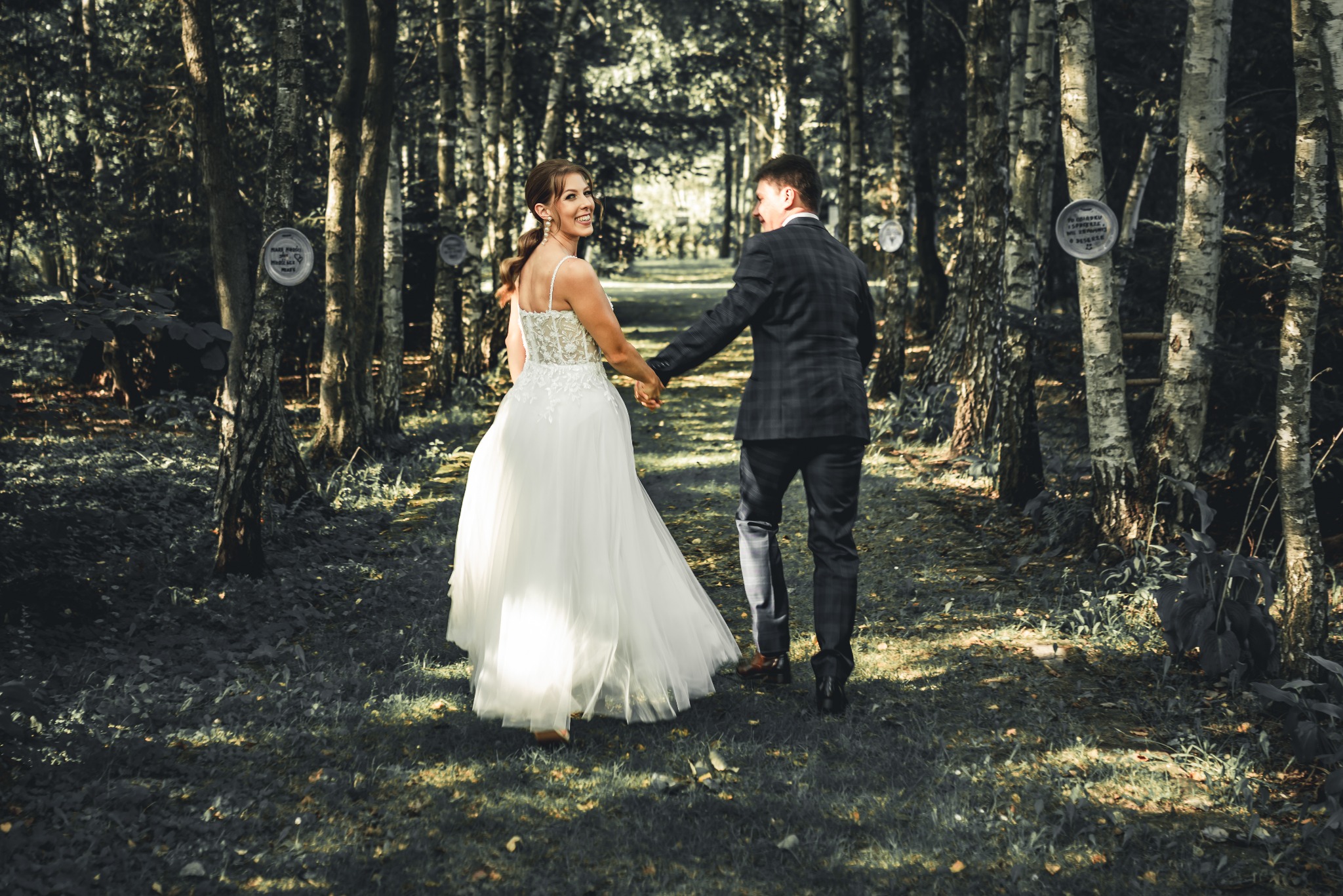 Para młoda idzie ścieżką w lesie, trzymając się za ręce. Panna młoda w białej sukni z koronkową górą spogląda przez ramię, uśmiechając się. Na drzewach widoczne okrągłe tabliczki z napisami.