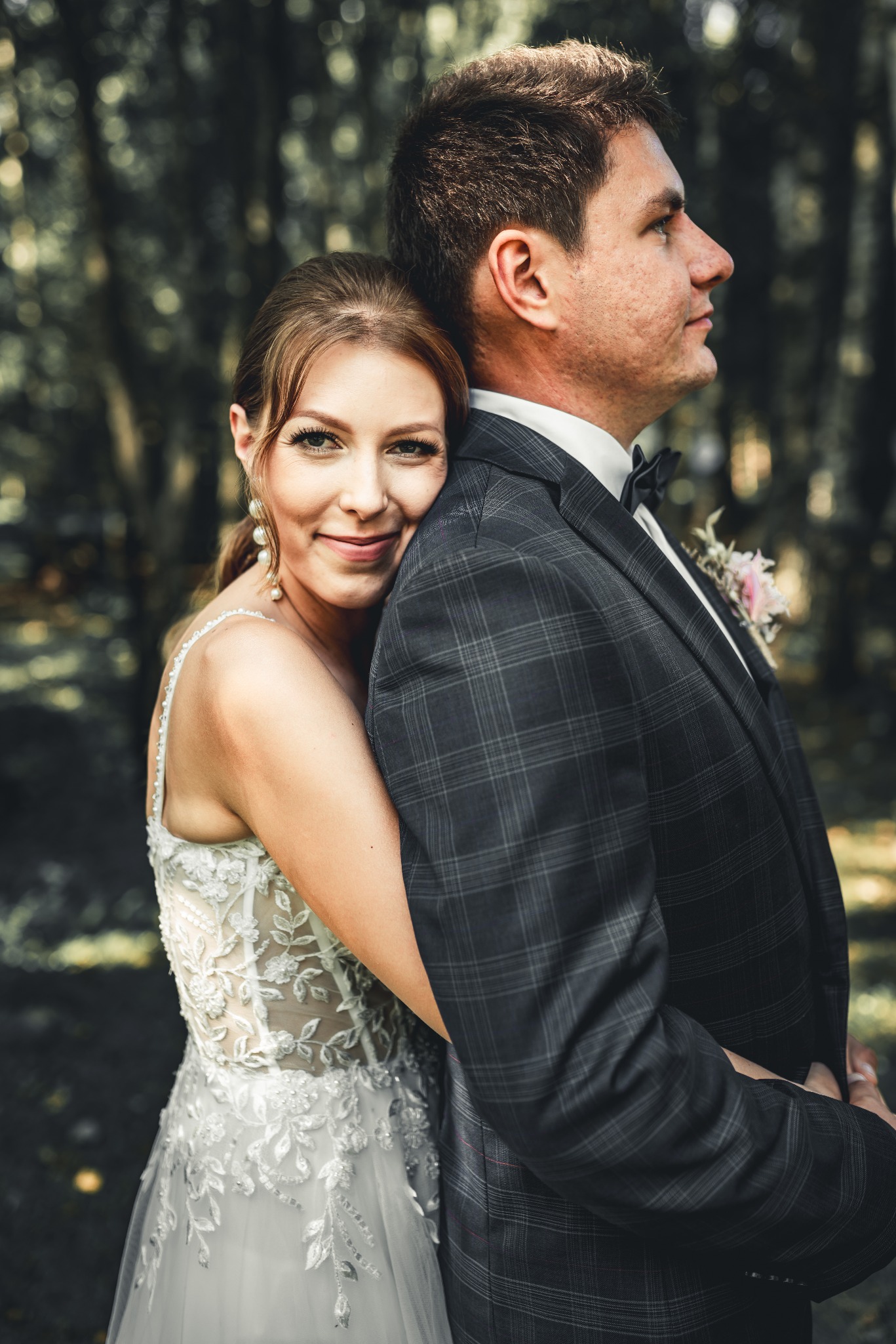 Panna młoda obejmuje pana młodego w ciemnoszarym garniturze w kratę, delikatny bukiecik w butonierce, ona w białej sukni z koronkowymi aplikacjami i perłowymi kolczykami, oboje uśmiechnięci na tle...