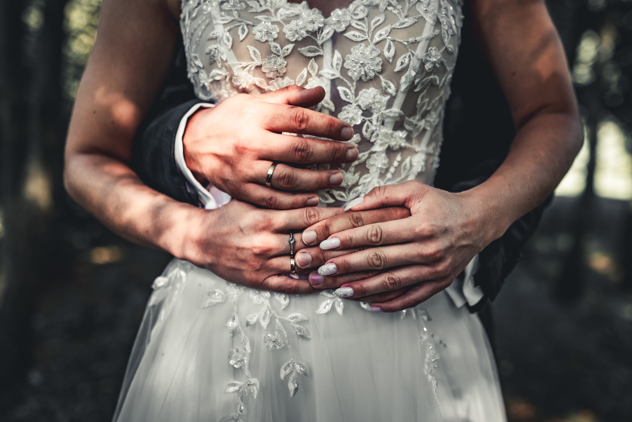 Dłonie mężczyzny obejmują kobietę w białej sukni ślubnej z koronkowymi aplikacjami, widoczne obrączki na palcach.