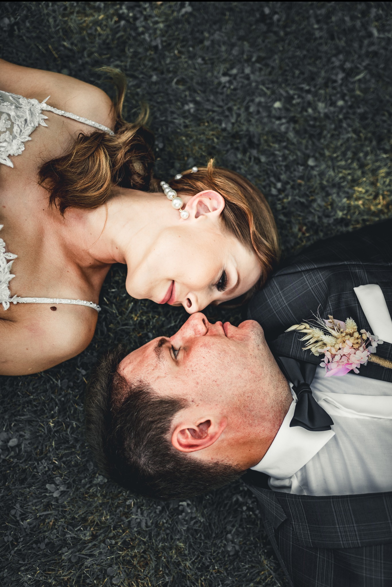 Para młoda leżąca na trawie, panna młoda w sukni z koronkową górą i perłowym naszyjnikiem, pan młody w garniturze w kratę z butonierką, zbliżają się do pocałunku.