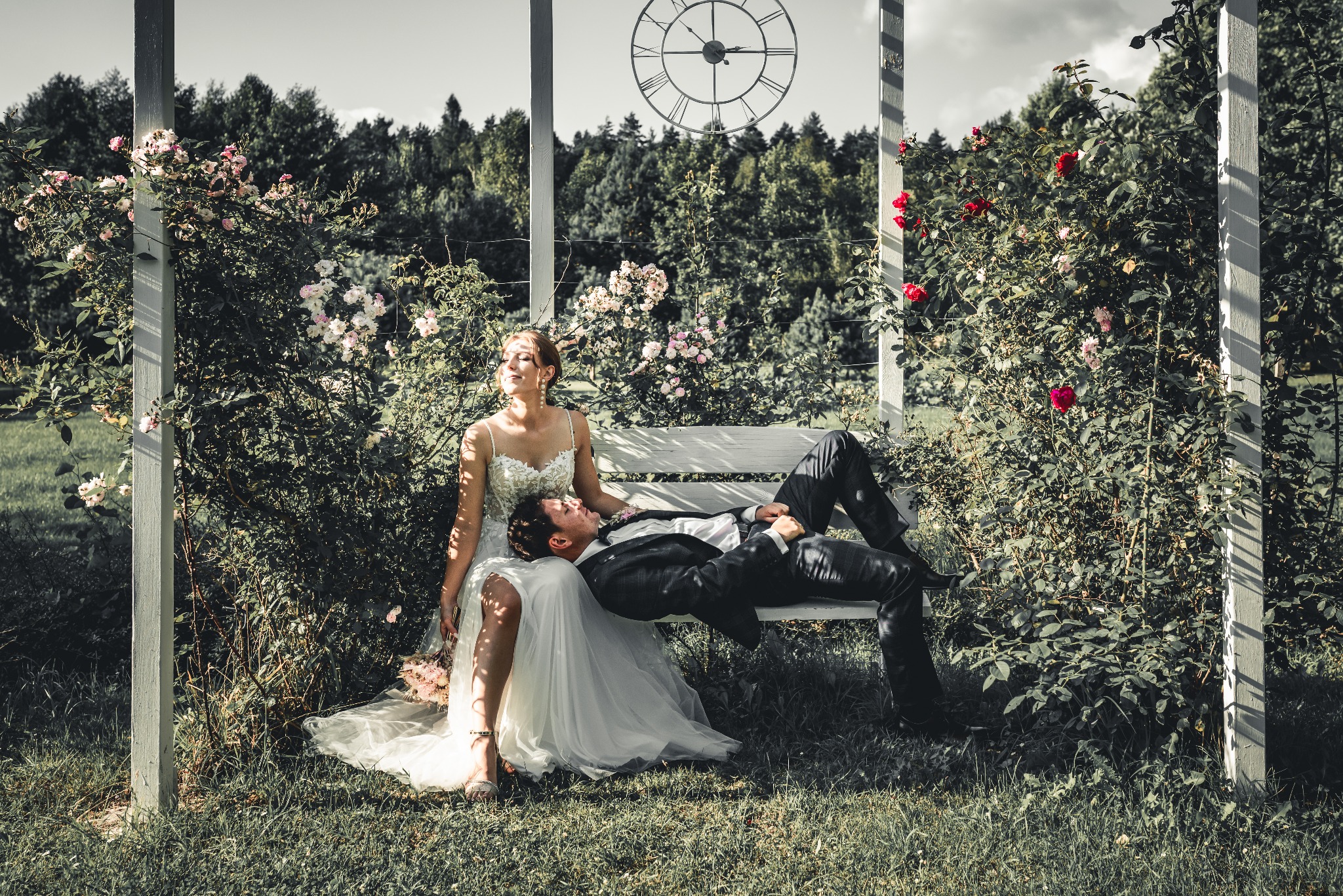 Para młoda odpoczywa na białej ławeczce otoczonej kwitnącymi różami, w tle las i dekoracyjny zegar słoneczny.