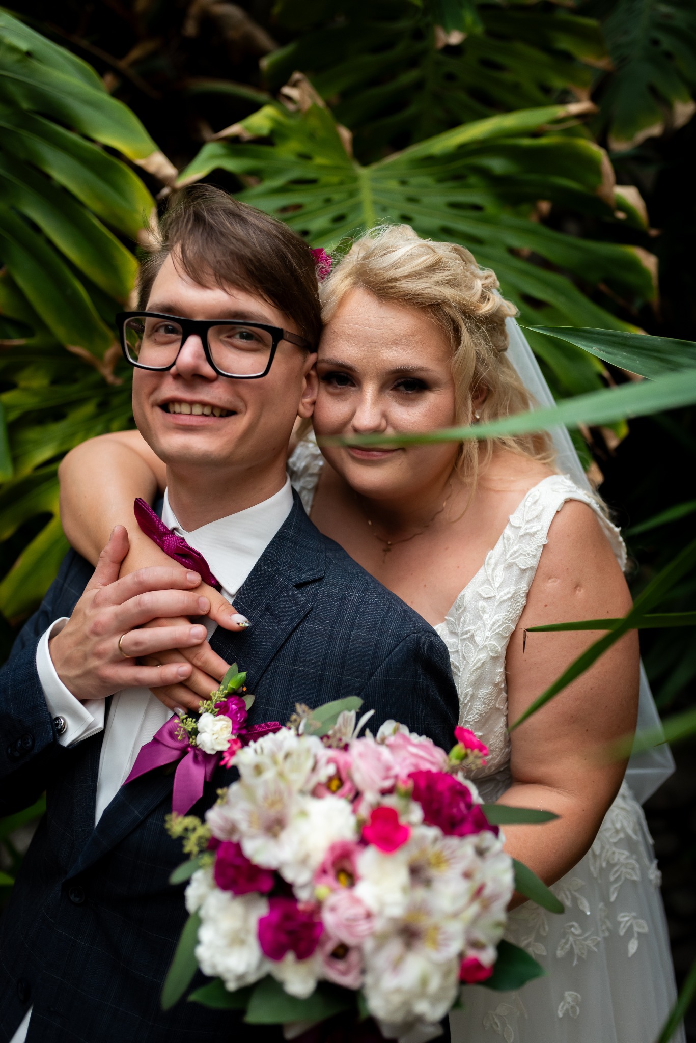 Uśmiechnięta para młoda w objęciach na tle tropikalnej zieleni; on w granatowym garniturze z bordową muchą, ona w białej sukni z koronkowymi detalami i welonem, trzymająca bukiet z różowych...