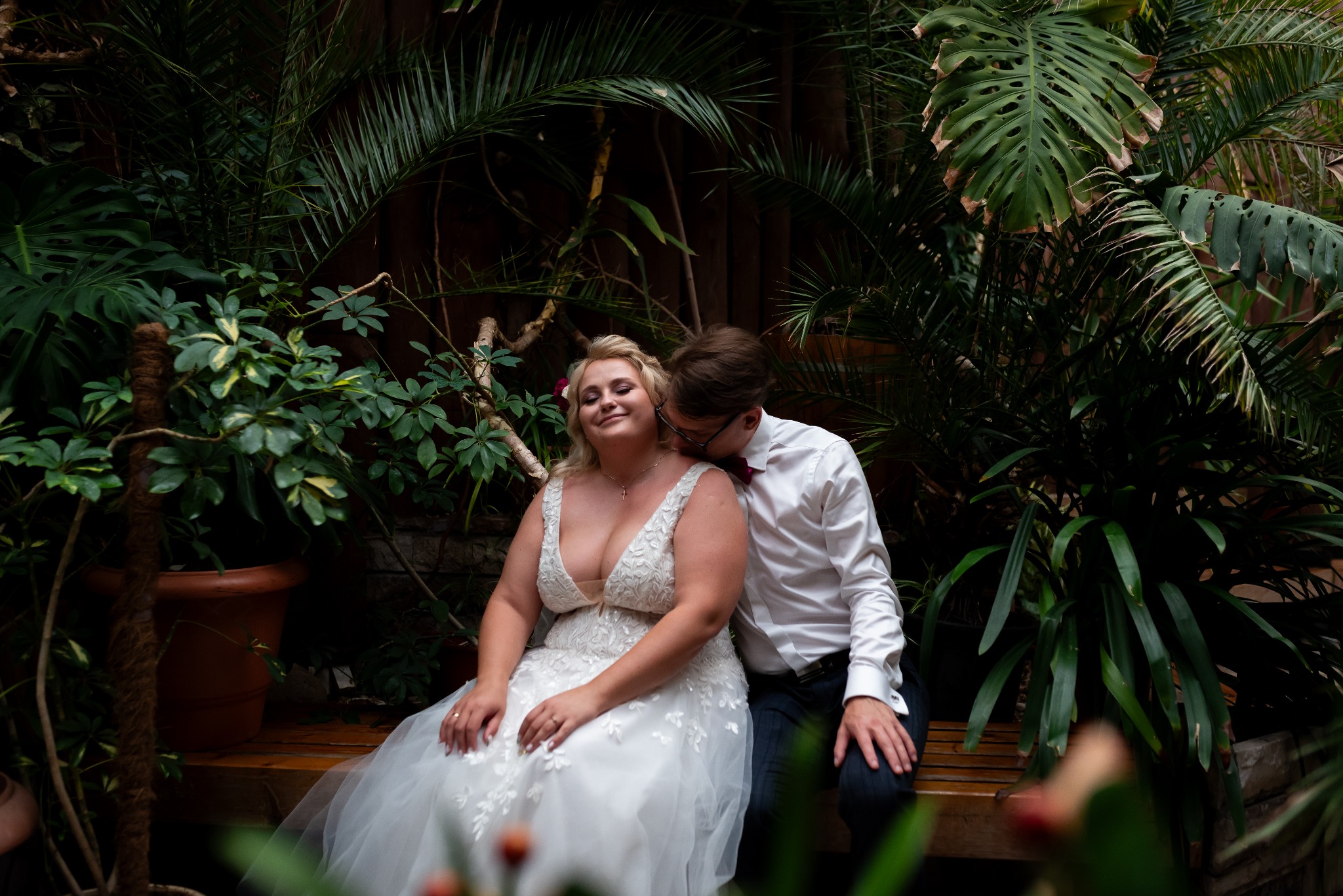 Para młoda w szklarni: Panna młoda w sukni ślubnej siedzi na ławce, oblubieniec w muszce całuje ją w szyję na tle bujnej zieleni.
