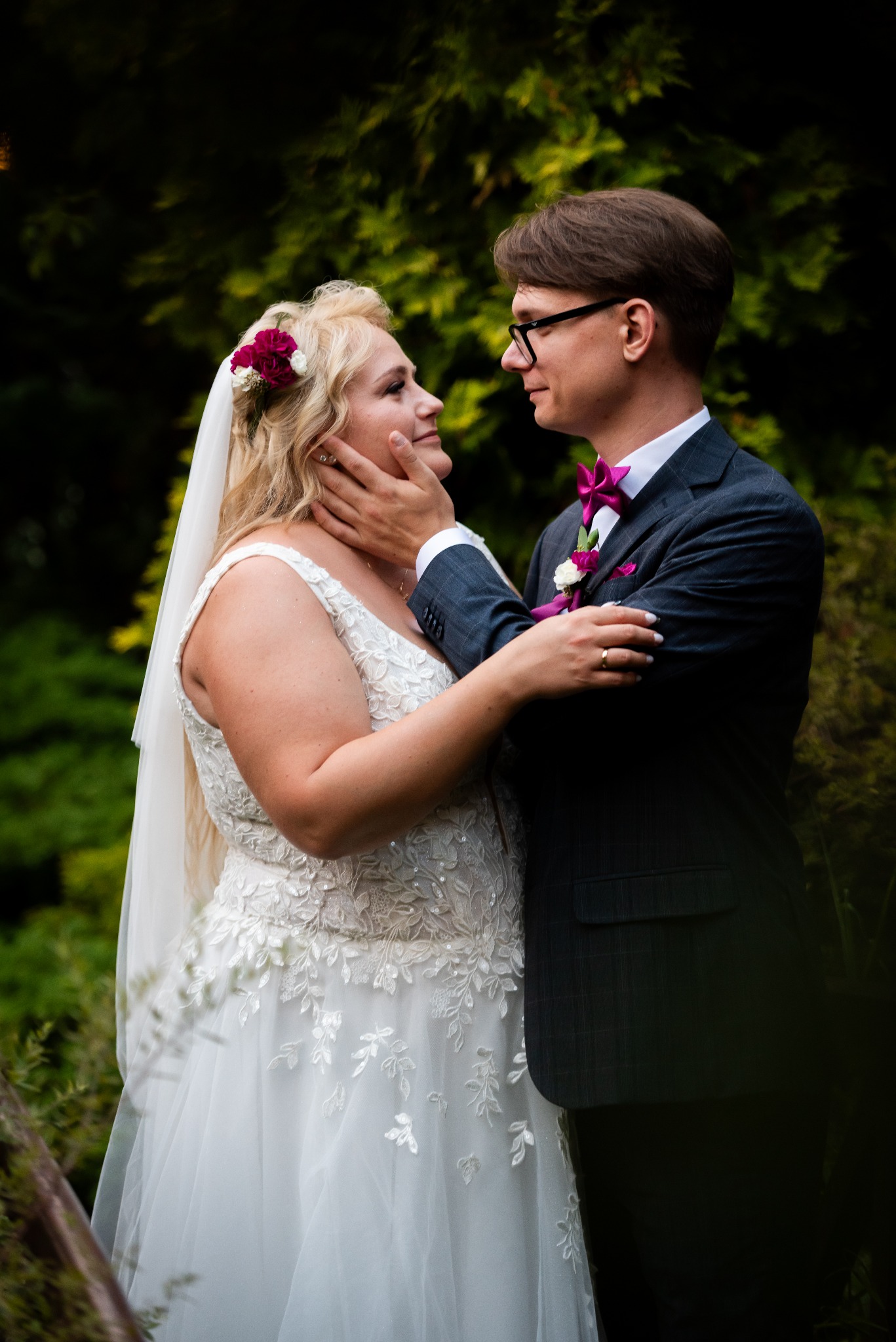 Para młoda: panna młoda w koronkowej sukni z kwiatami we włosach, pan młody w ciemnym garniturze z bordową muchą, patrzą na siebie z czułością na tle zieleni.