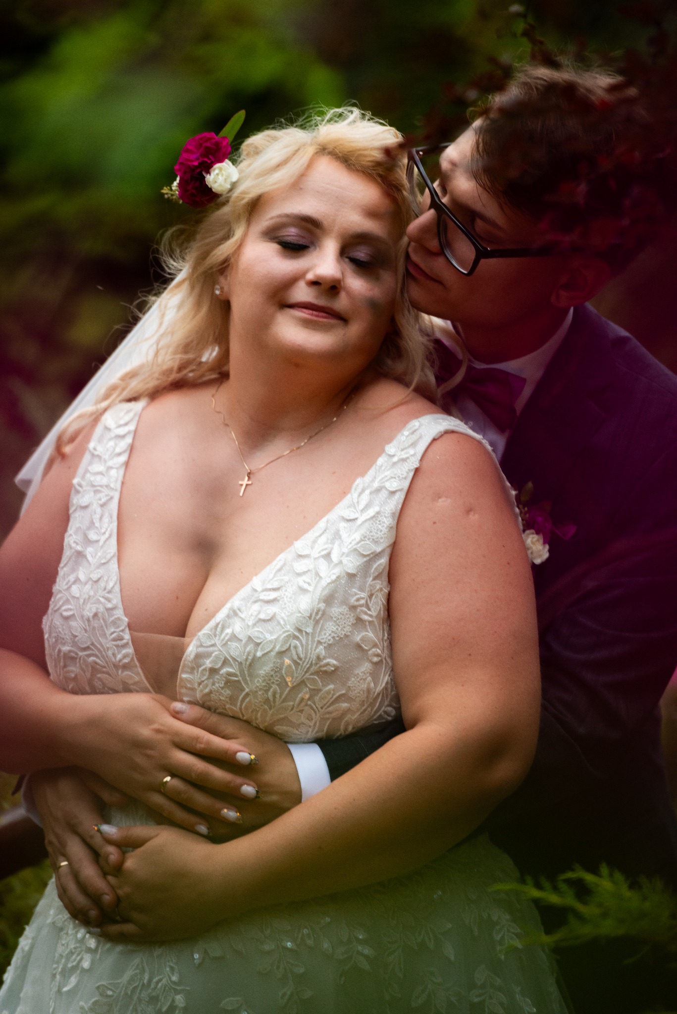 Uśmiechnięta panna młoda w koronkowej sukni z kwiatami we włosach, obejmowana przez pana młodego w okularach i bordowej muszce, który ją całuje.