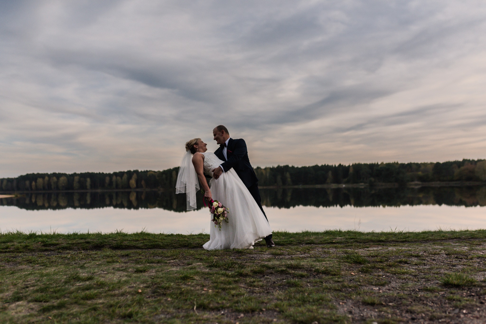 Para młoda w tańcu na tle jeziora i pochmurnego nieba, panna młoda w białej sukni z bukietem, pan młody w ciemnym garniturze.