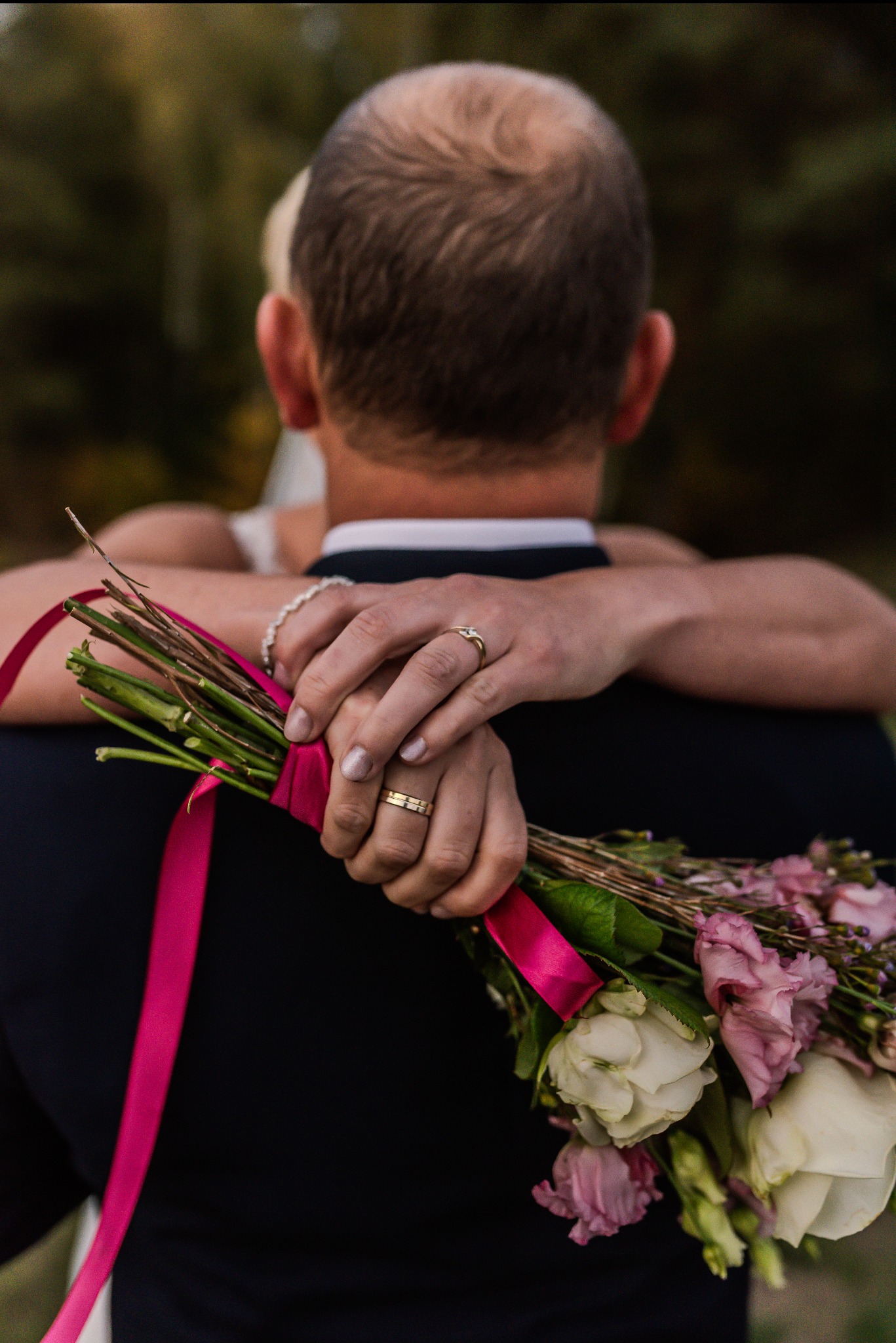 Para młoda obejmująca się, widoczne dłonie z obrączkami i bukiet ślubny z różową wstążką, perspektywa od tyłu.