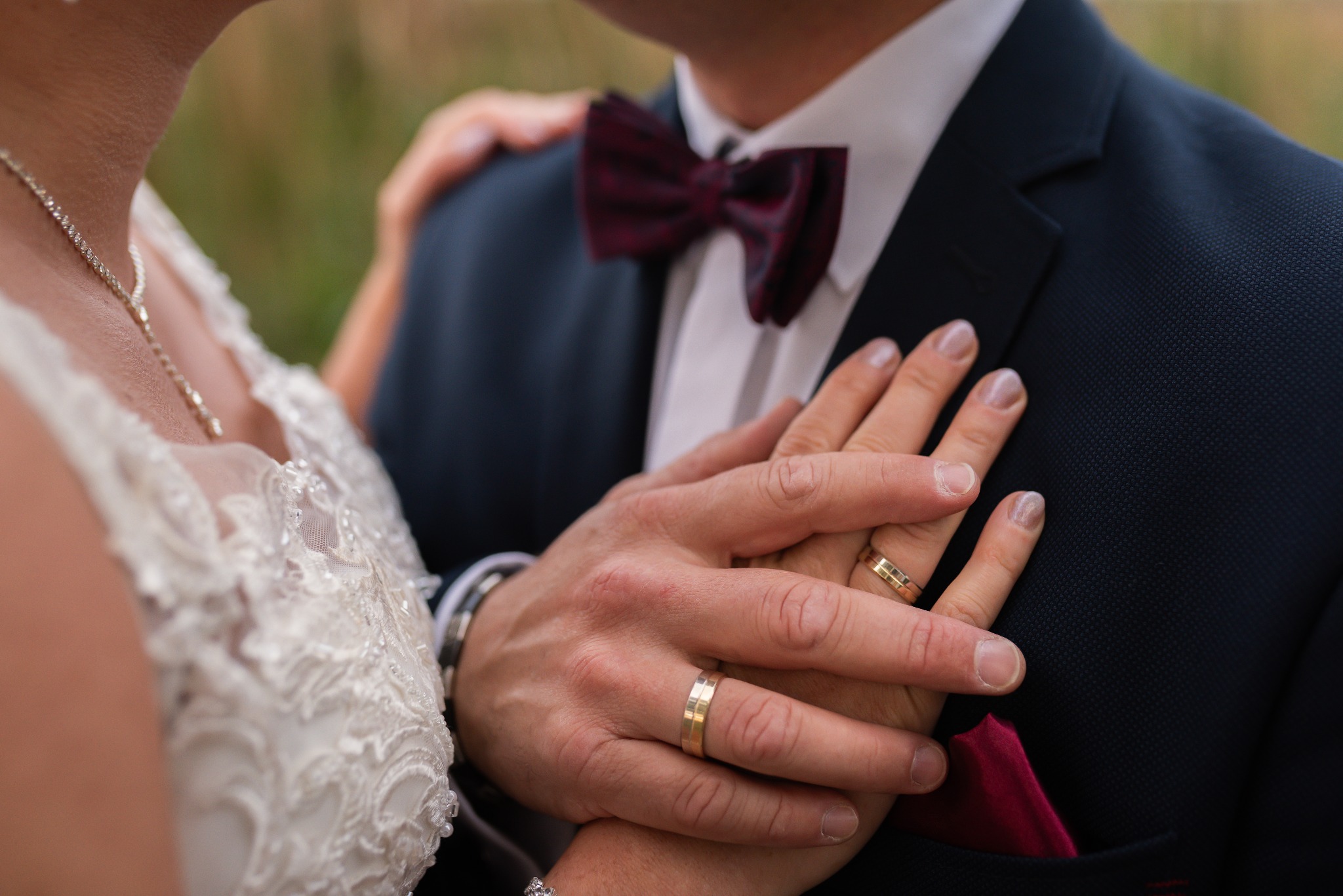 Dłonie pary młodej z obrączkami splecione na tle granatowego garnituru z bordową muchą i białej sukni z koronkowymi zdobieniami.