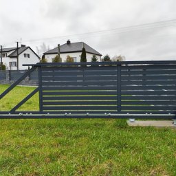 AK TECHNIK ALBERT KOBRZENIECKI - Nowoczesna, przesuwna brama wjazdowa z automatyką, szara, poziome panele, na tle trawnika i domów jednorodzinnych w pochmurny dzień. Widoczny mechanizm bramy.