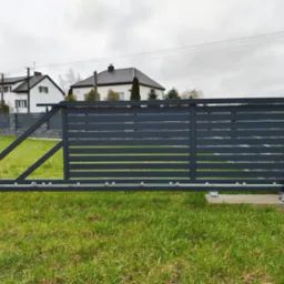 Nowoczesna, przesuwna brama wjazdowa z automatyką, szara, poziome panele, na tle trawnika i domów jednorodzinnych w pochmurny dzień. Widoczny mechanizm bramy.