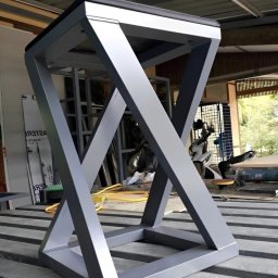 AK TECHNIK ALBERT KOBRZENIECKI - Metalowa konstrukcja w kształcie sześcianu z ukośnymi wzmocnieniami, pomalowana na szaro, stoi na metalowych profilach. Widoczny warsztat w tle.