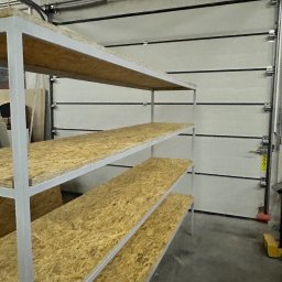AK TECHNIK ALBERT KOBRZENIECKI - Warsztat: metalowy regał z płytami OSB na tle segmentowej bramy garażowej z widocznymi elementami montażowymi i instalacjami pod sufitem.