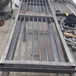 AK TECHNIK ALBERT KOBRZENIECKI - Spawana konstrukcja metalowa z rurkami w kolorze niebiesko-czerwonym, leżąca na stole warsztatowym. Widoczne narzędzia i akcesoria spawalnicze w tle.