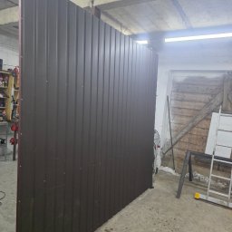 AK TECHNIK ALBERT KOBRZENIECKI - Ciemnobrązowe ogrodzenie panelowe z widocznymi śrubami mocującymi, ustawione w surowym wnętrzu warsztatu z betonową podłogą i drewnianymi elementami w tle.