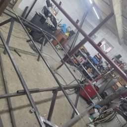 AK TECHNIK ALBERT KOBRZENIECKI - Konstrukcja metalowa w warsztacie, spawana rama z profili stalowych, widoczna spawarka i narzędzia, surowe wykończenie, szlifowane spawy, przygotowanie do dalszej obróbki.