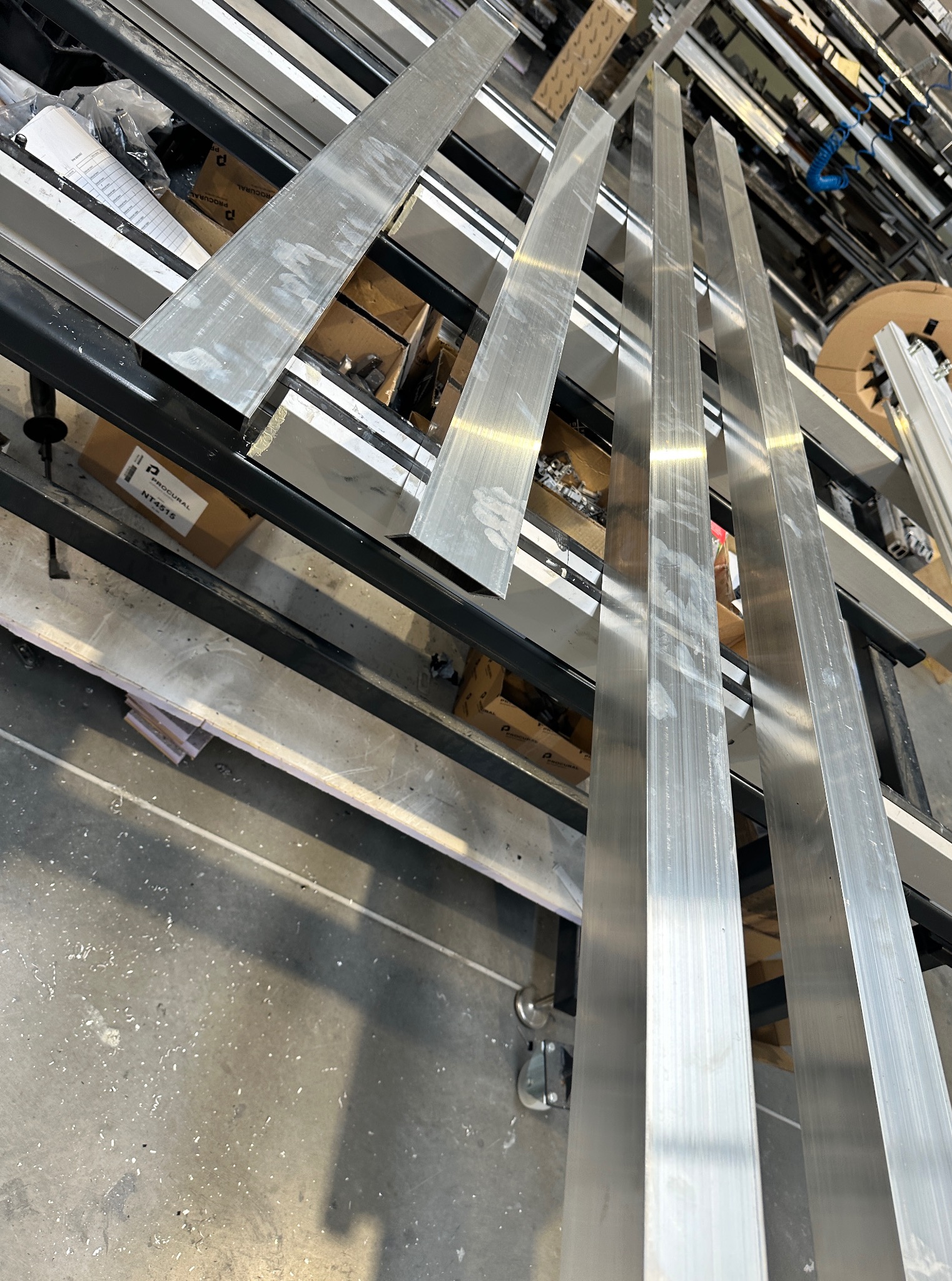 Przygotowane profile aluminiowe na regale warsztatowym, gotowe do obróbki lub montażu. Widoczne detale powierzchni i otoczenia warsztatowego.