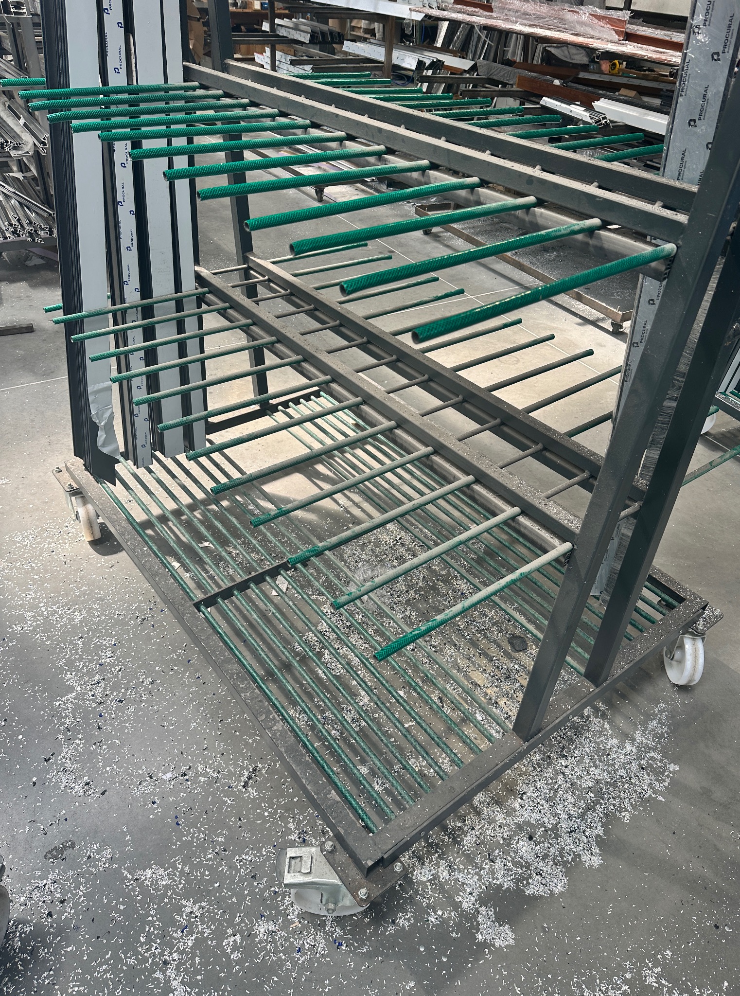 Wózek warsztatowy z metalową konstrukcją i zielonymi poprzeczkami, na którym składowane są aluminiowe profile. W tle widoczne inne elementy i odpady po obróbce.