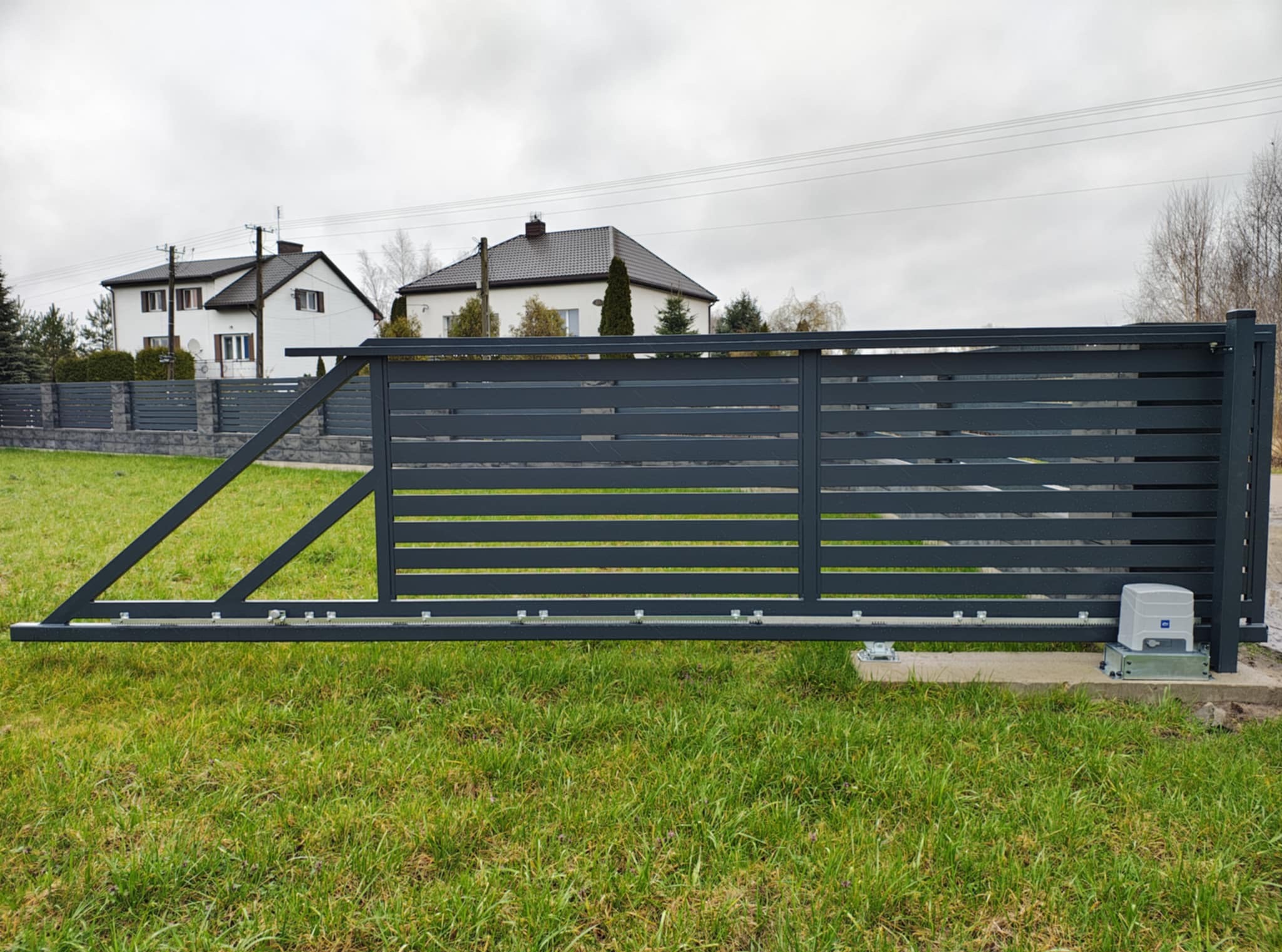 Nowoczesna, przesuwna brama wjazdowa z automatyką, szara, poziome panele, na tle trawnika i domów jednorodzinnych w pochmurny dzień. Widoczny mechanizm bramy.