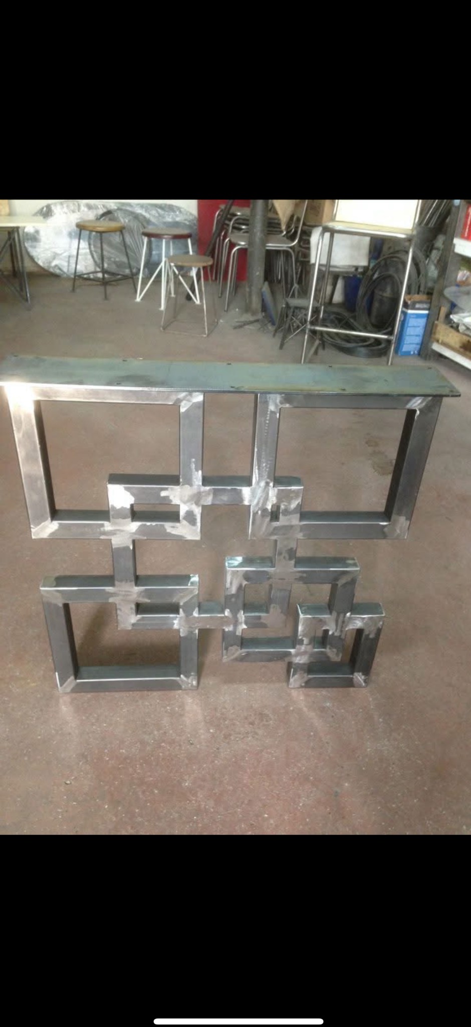 Spawana konstrukcja metalowa z kwadratów, surowy metal, widoczne spawy, tło warsztatowe z krzesłami i stołami. Konstrukcja prawdopodobnie do balustrady.