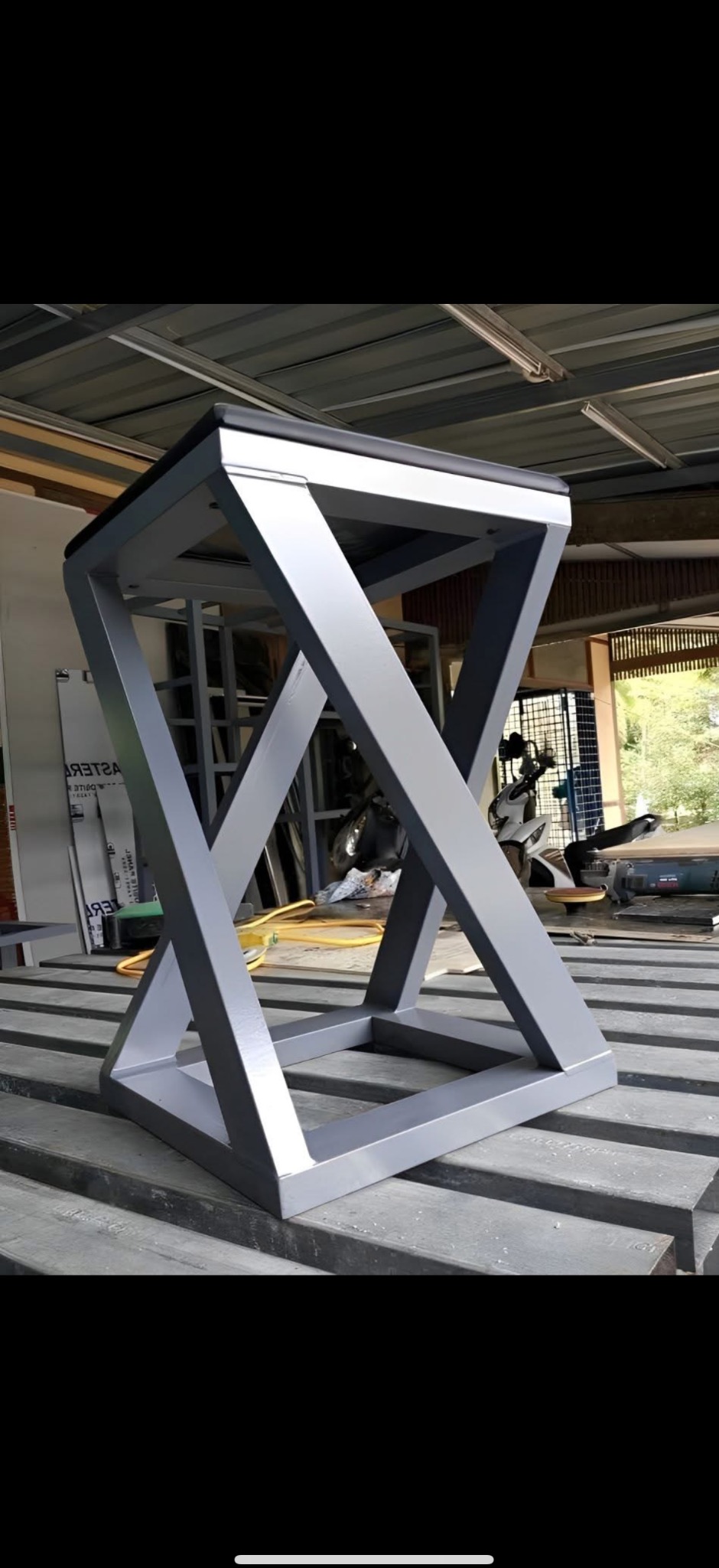 Metalowa konstrukcja w kształcie sześcianu z ukośnymi wzmocnieniami, pomalowana na szaro, stoi na metalowych profilach. Widoczny warsztat w tle.