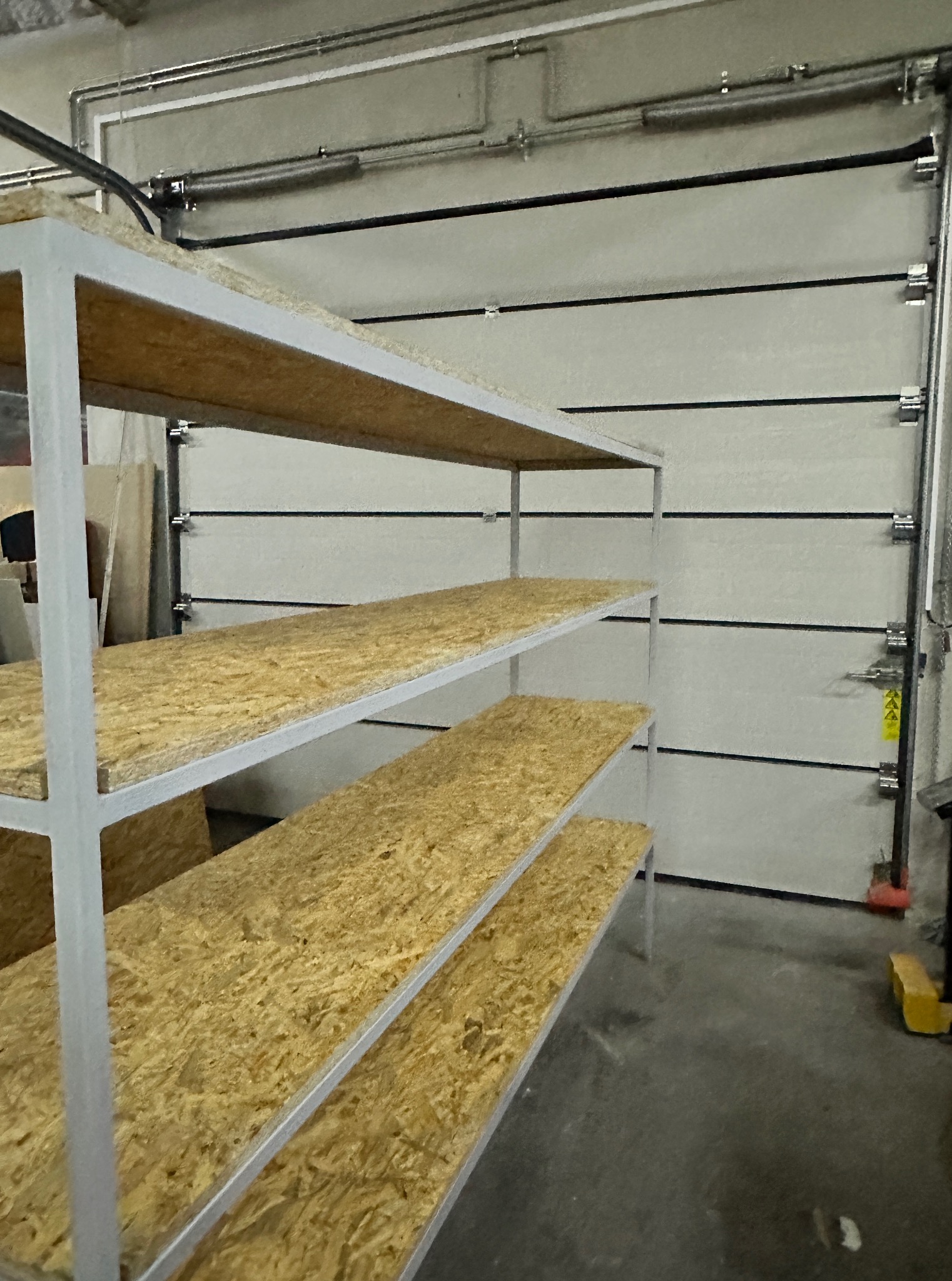 Warsztat: metalowy regał z płytami OSB na tle segmentowej bramy garażowej z widocznymi elementami montażowymi i instalacjami pod sufitem.