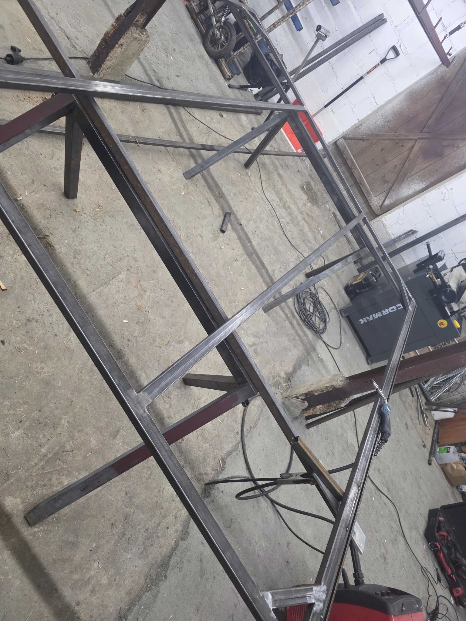 Metalowa konstrukcja w trakcie spawania, widoczne spawy i narzędzia w warsztacie. Surowy, industrialny charakter.