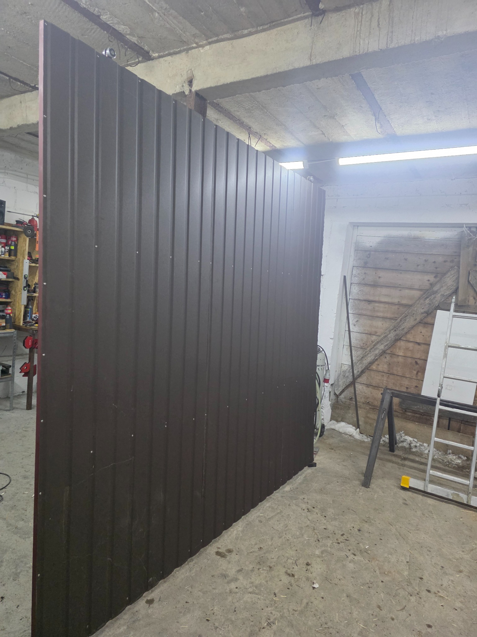 Ciemnobrązowe ogrodzenie panelowe w trakcie montażu w Czachy-Kołaki. Widoczne mocowania i surowy beton posadzki. Częściowo widoczna drabina i drewniane elementy.