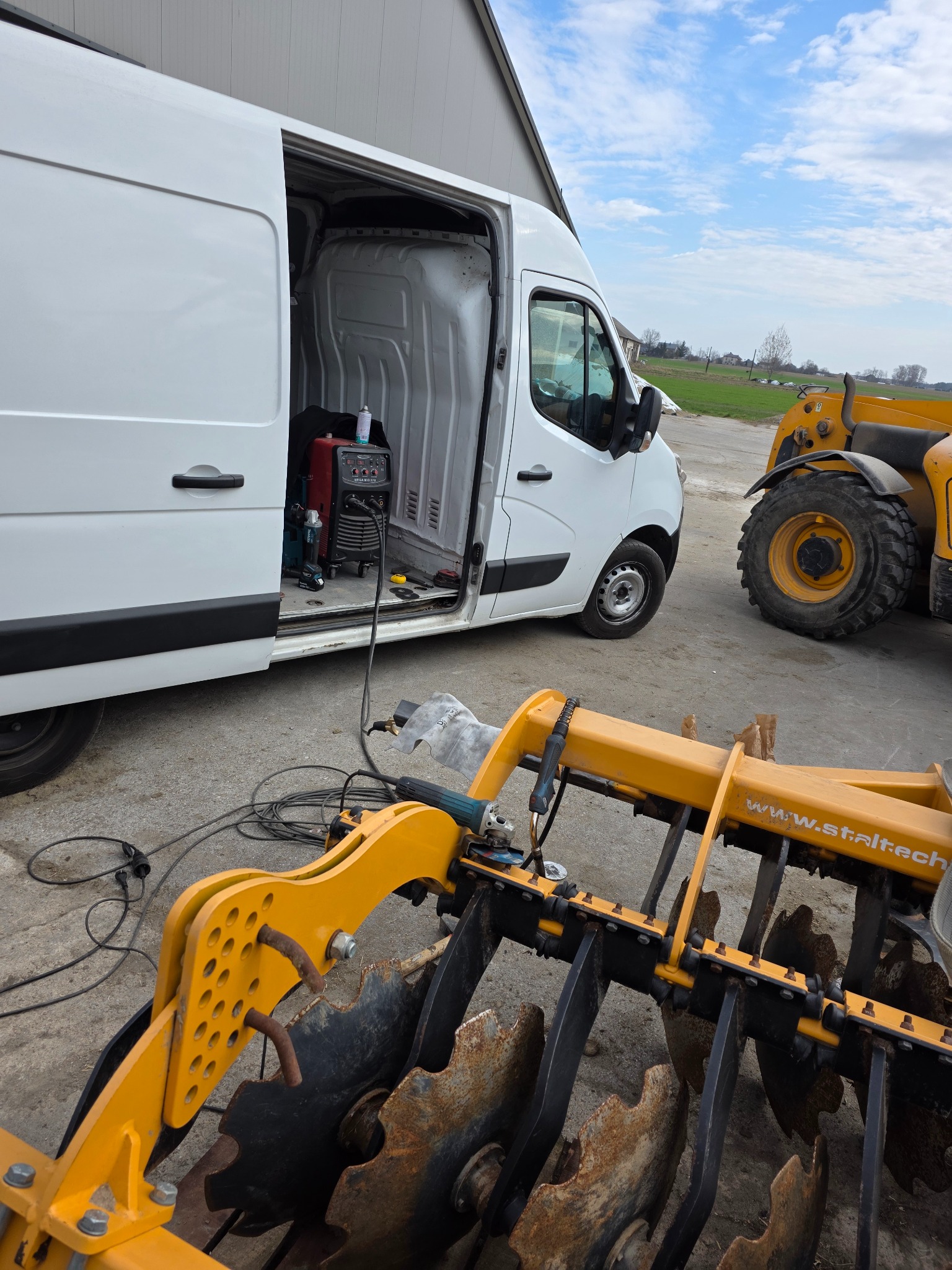 Mobilny spawacz przy pracy: żółty agregat rolniczy, spawarka w busie. Sprzęt na zewnątrz, w tle budynek i niebo. Widoczne przewody i narzędzia.