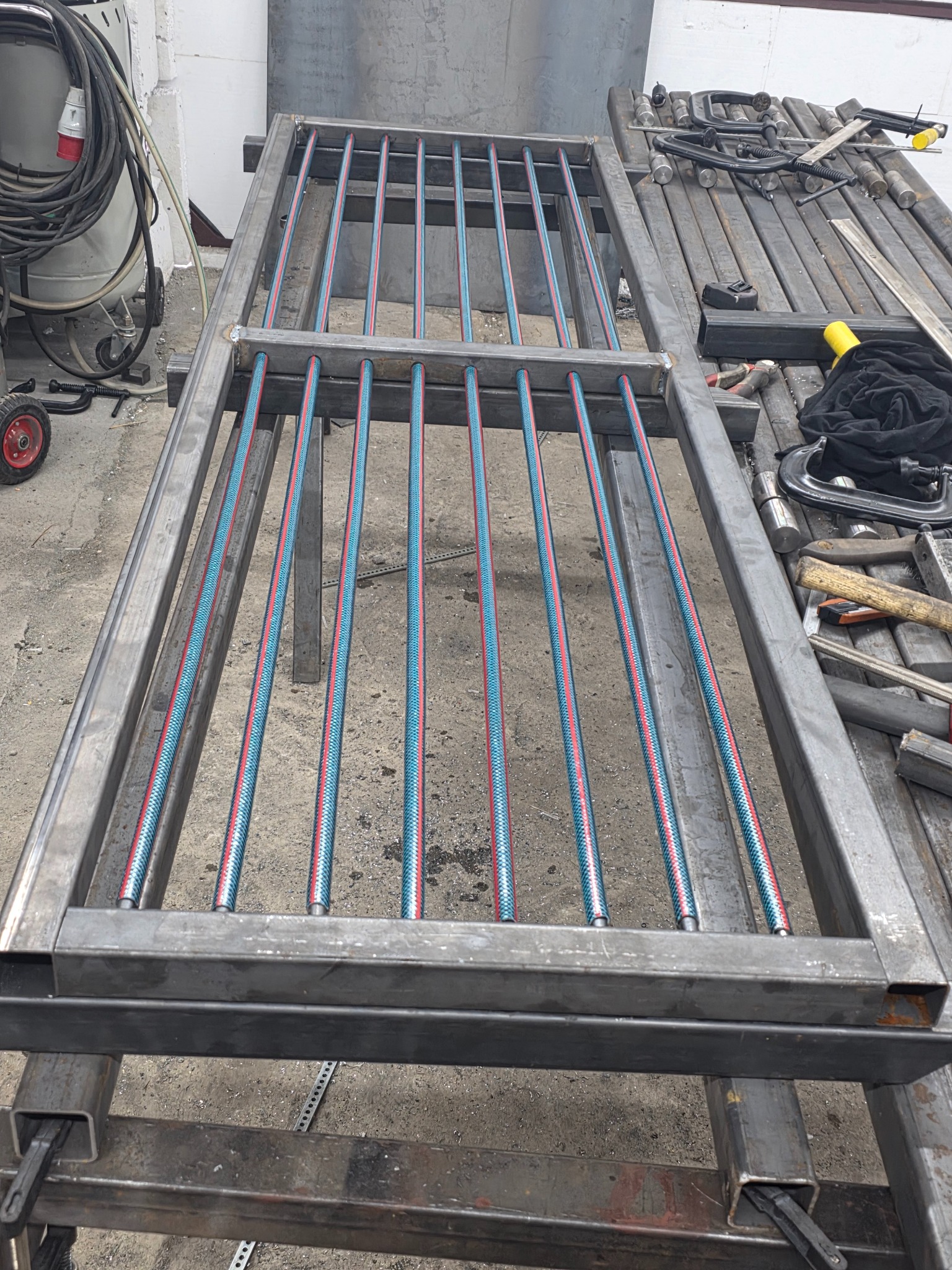 Spawana konstrukcja metalowa z rurkami w kolorze niebiesko-czerwonym, leżąca na stole warsztatowym. Widoczne narzędzia i akcesoria spawalnicze w tle.