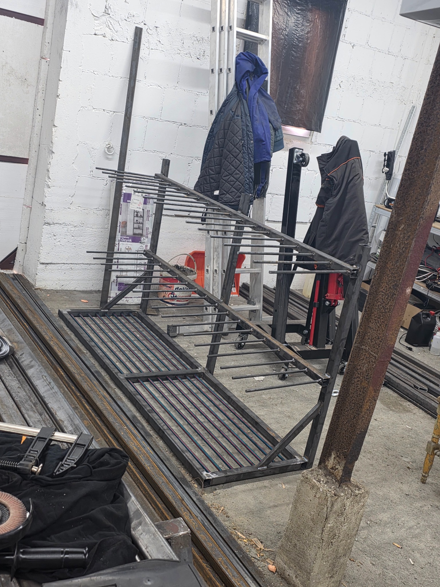 W warsztacie: konstrukcja metalowa z prętami, prawdopodobnie balustrada lub element ogrodzenia, w trakcie spawania. Widoczne narzędzia i materiały.