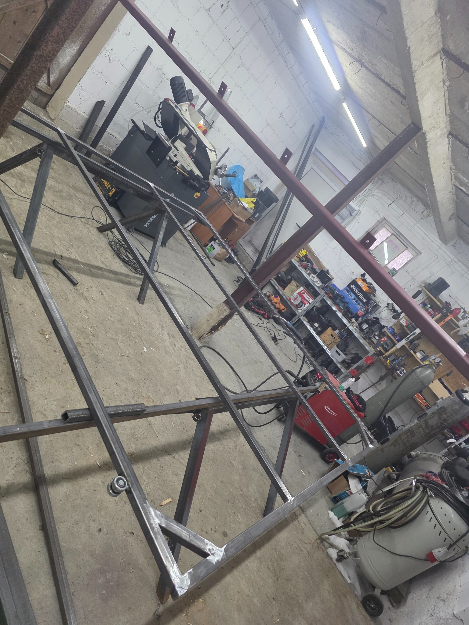 Konstrukcja metalowa w warsztacie, spawana rama z profili stalowych, widoczna spawarka i narzędzia, surowe wykończenie, szlifowane spawy, przygotowanie do dalszej obróbki.