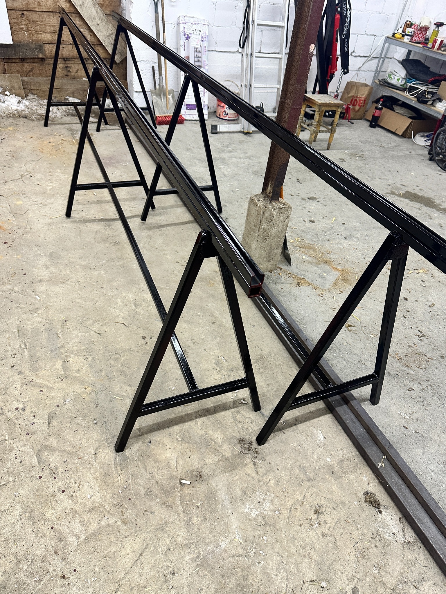 Czarne, metalowe kozły spawalnicze podtrzymujące długi profil. Widoczne ślady spawania i obróbki. Surowe, warsztatowe otoczenie.