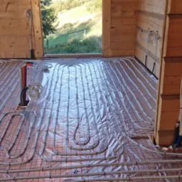 Montaż rur grzewczych podłogowych na folii odblaskowej z siatką w drewnianym domu w trakcie budowy, widoczne przewody i drabina.